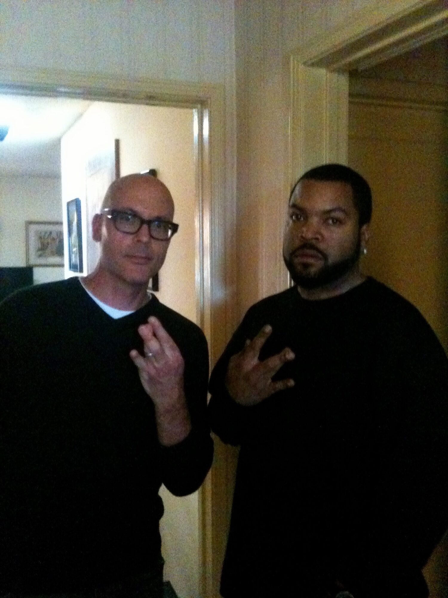 Two men standing indoors, both making hand gestures, with a doorway and wall visible behind them.