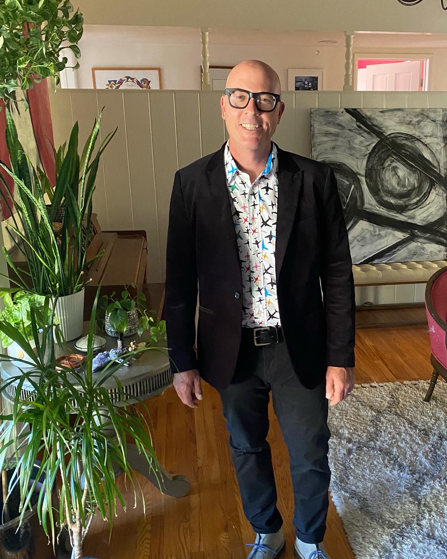 Man with glasses in a black blazer and patterned shirt standing indoors near a large plant and artwork.