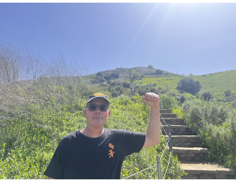 Person wearing sunglasses and a black shirt with a small orange logo, standing outdoors with a hill and stairs in the background.