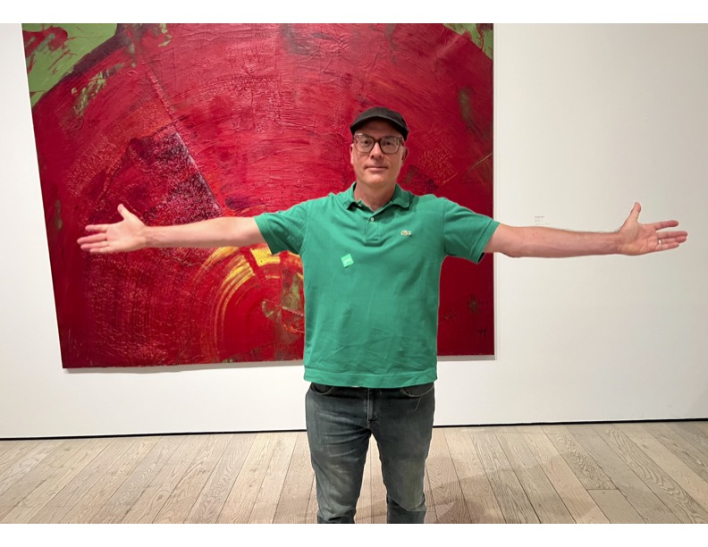Person standing with arms outstretched in front of a large red abstract painting, wearing a green shirt and glasses.