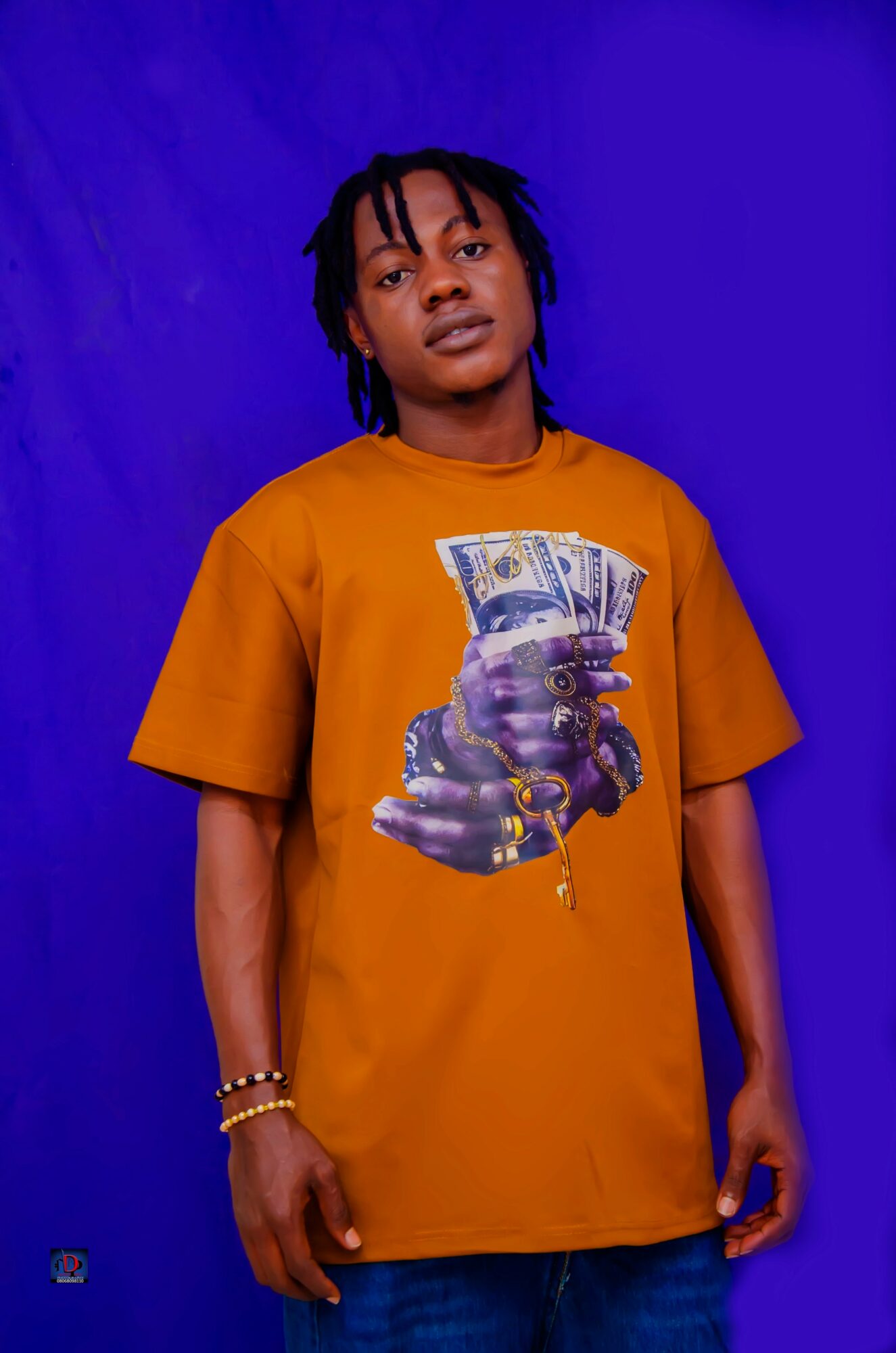 Young person with dreadlocks wearing an orange T-shirt with a graphic of a hand holding money, standing against a blue background.