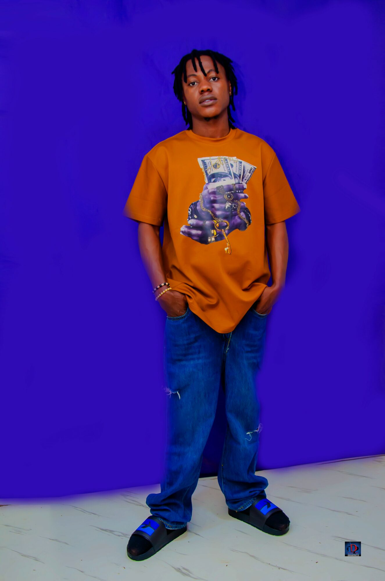 Young person with dreadlocks wearing an orange T-shirt with a money graphic, blue jeans, and sneakers, standing against a blue background.