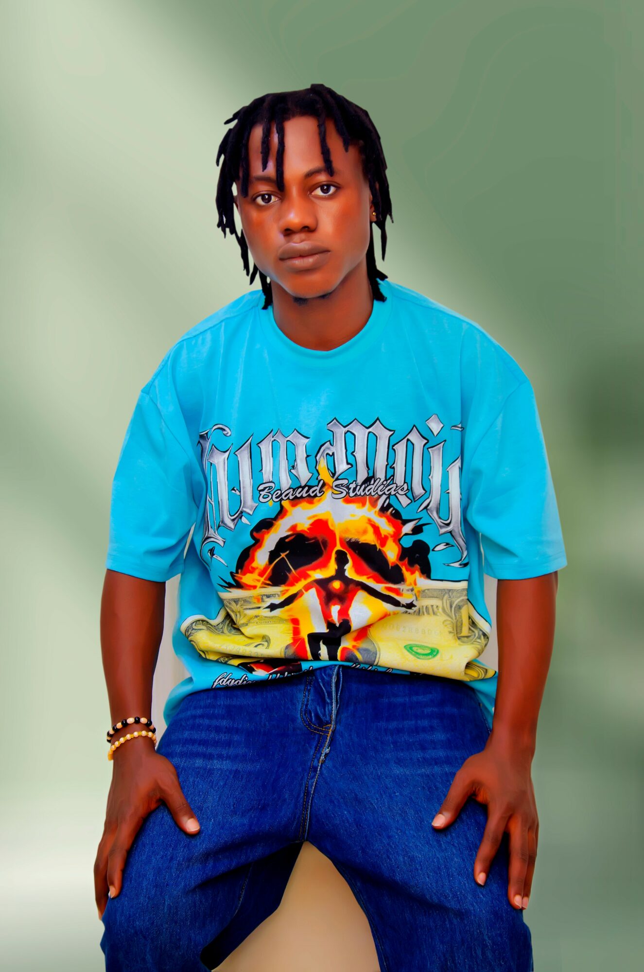 Young person with dreadlocks wearing a bright blue graphic T-shirt and loose jeans, standing against a green background.