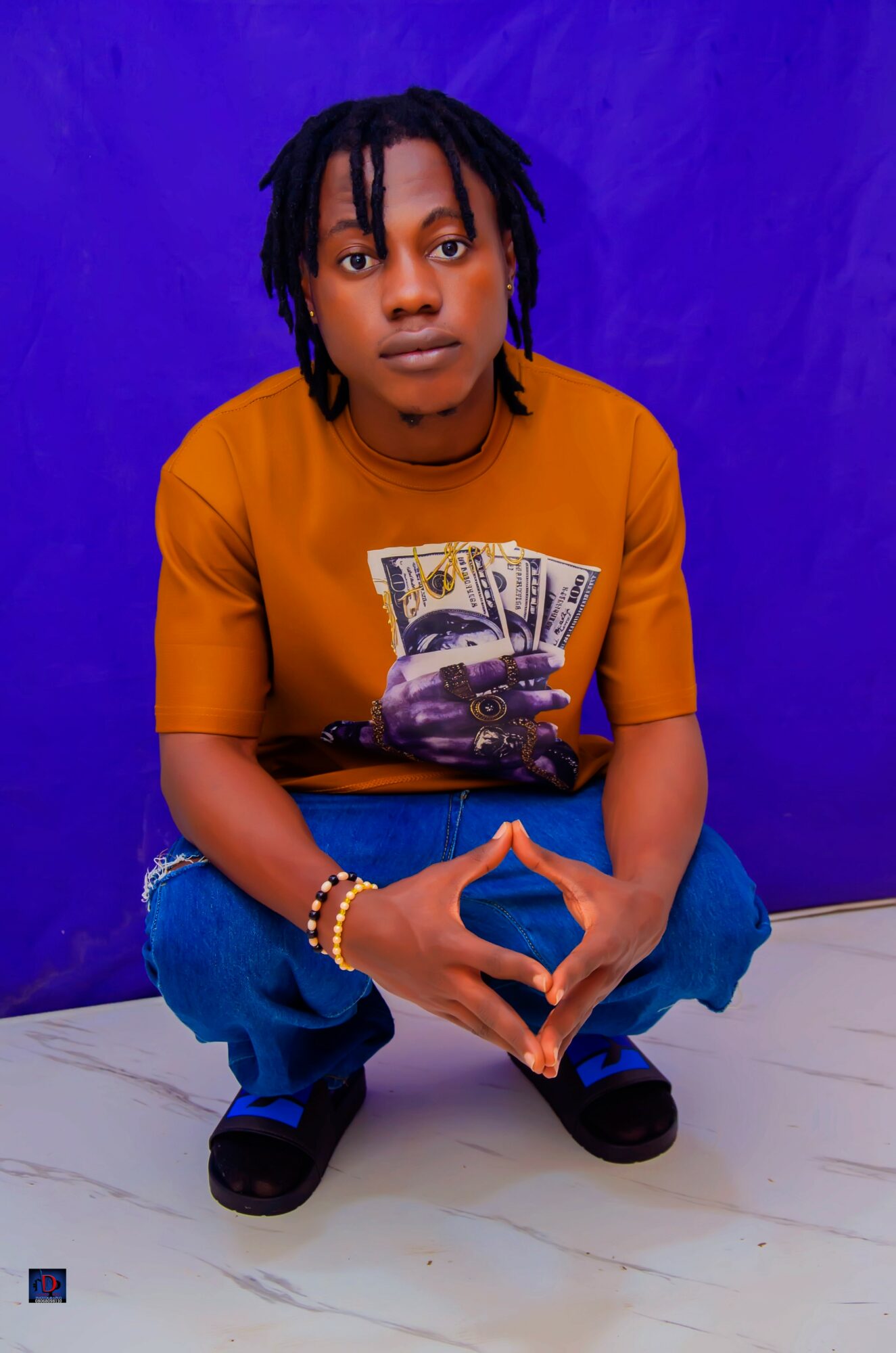 Person with dreadlocks crouching against a purple background, wearing a mustard yellow T-shirt with a dollar bill graphic, blue jeans, and black shoes.
