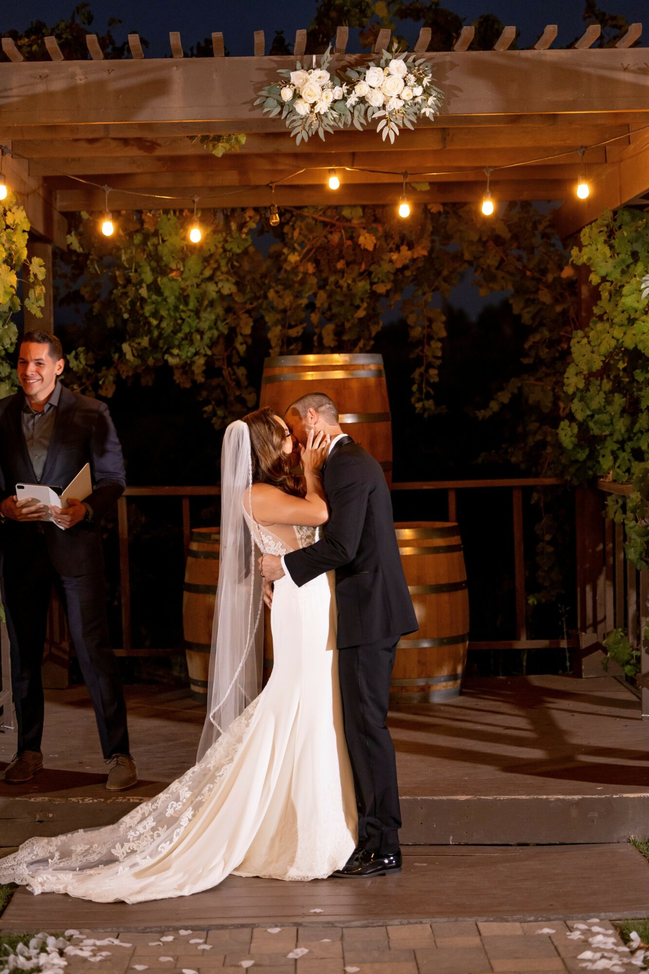 Bride and groom kiss under a decorated outdoor wooden pergola at night, with string lights and greenery, during a wedding ceremony.