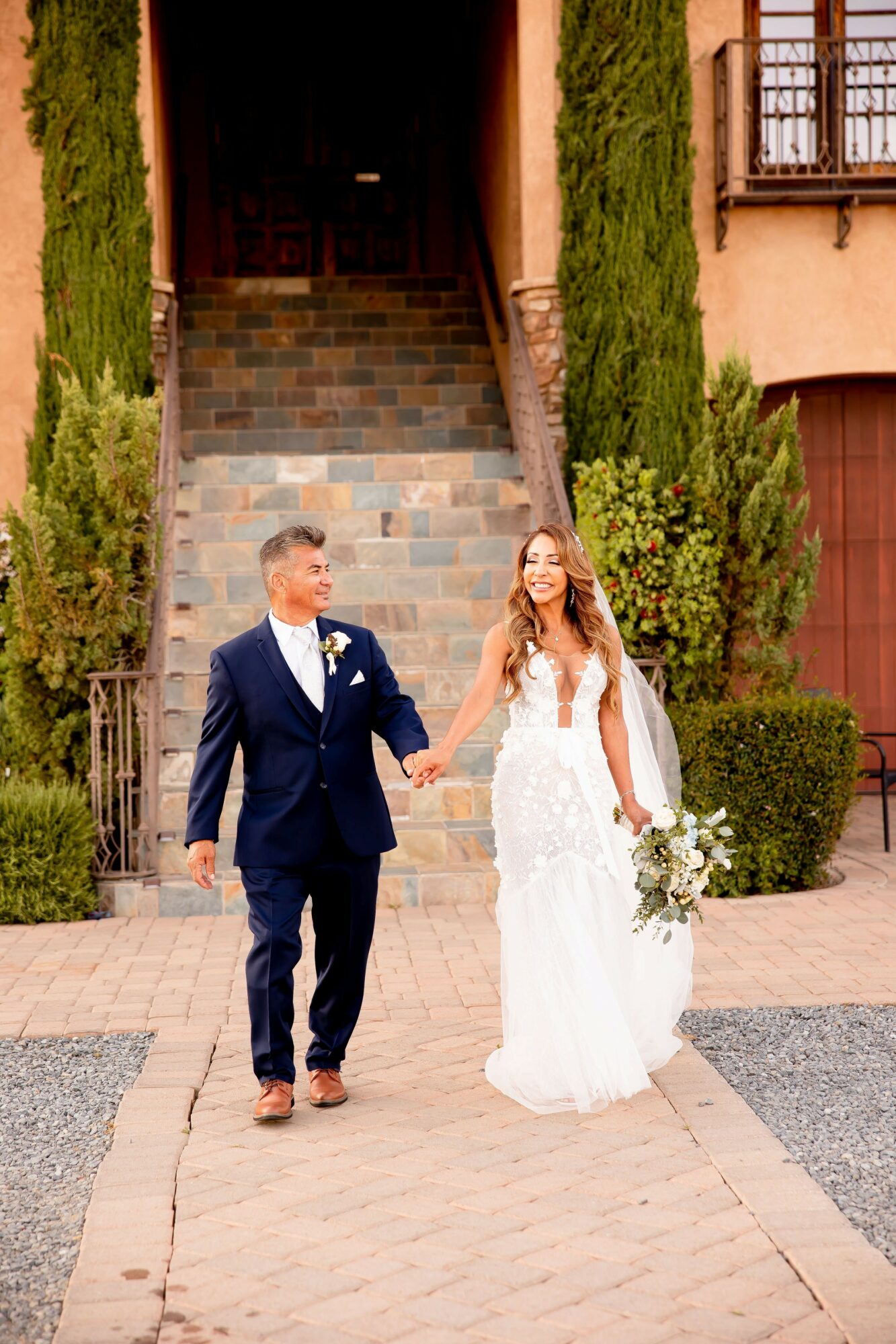 A bride and groom walk hand in hand outside a building with stairs, greenery, and a window, holding a bouquet.