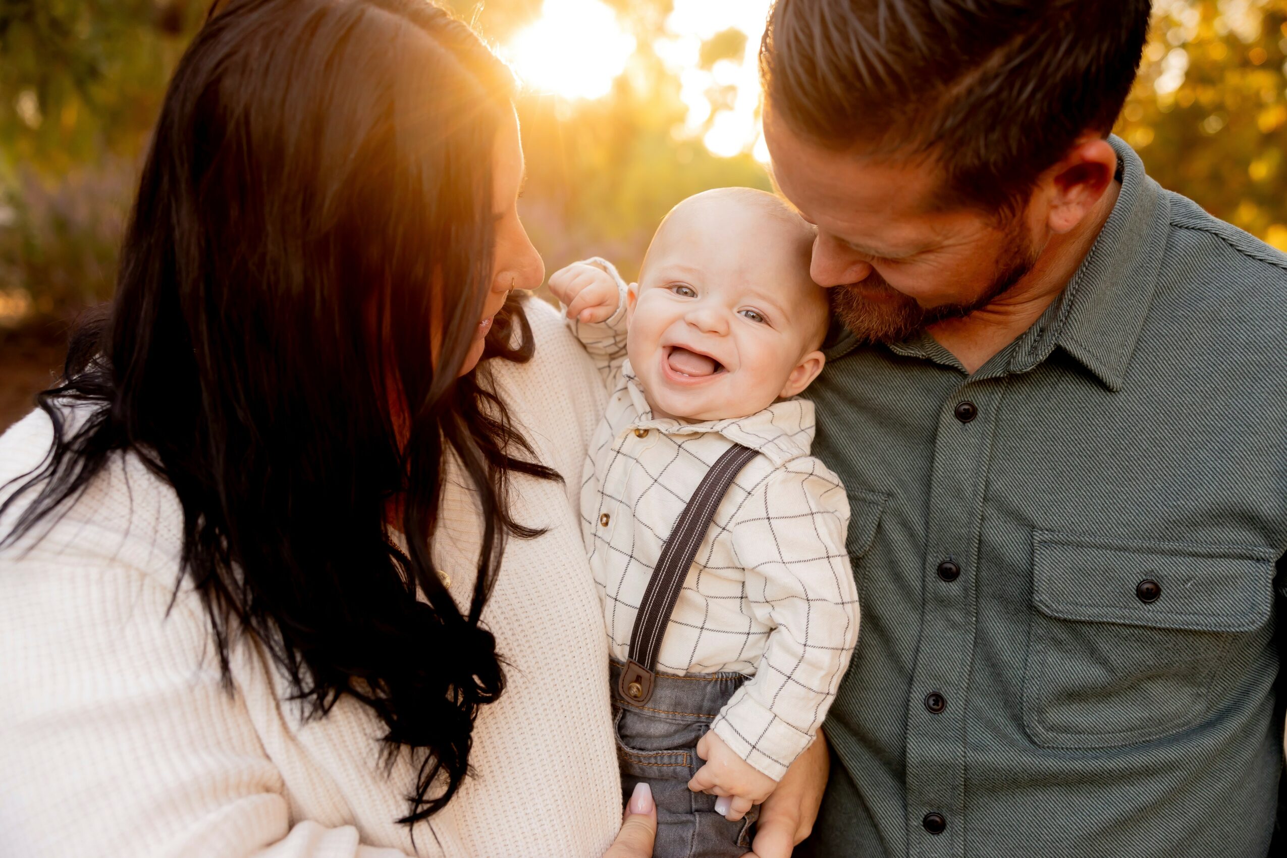 Family of three outdoors, smiling and looking at each other, with sunlight in the background.