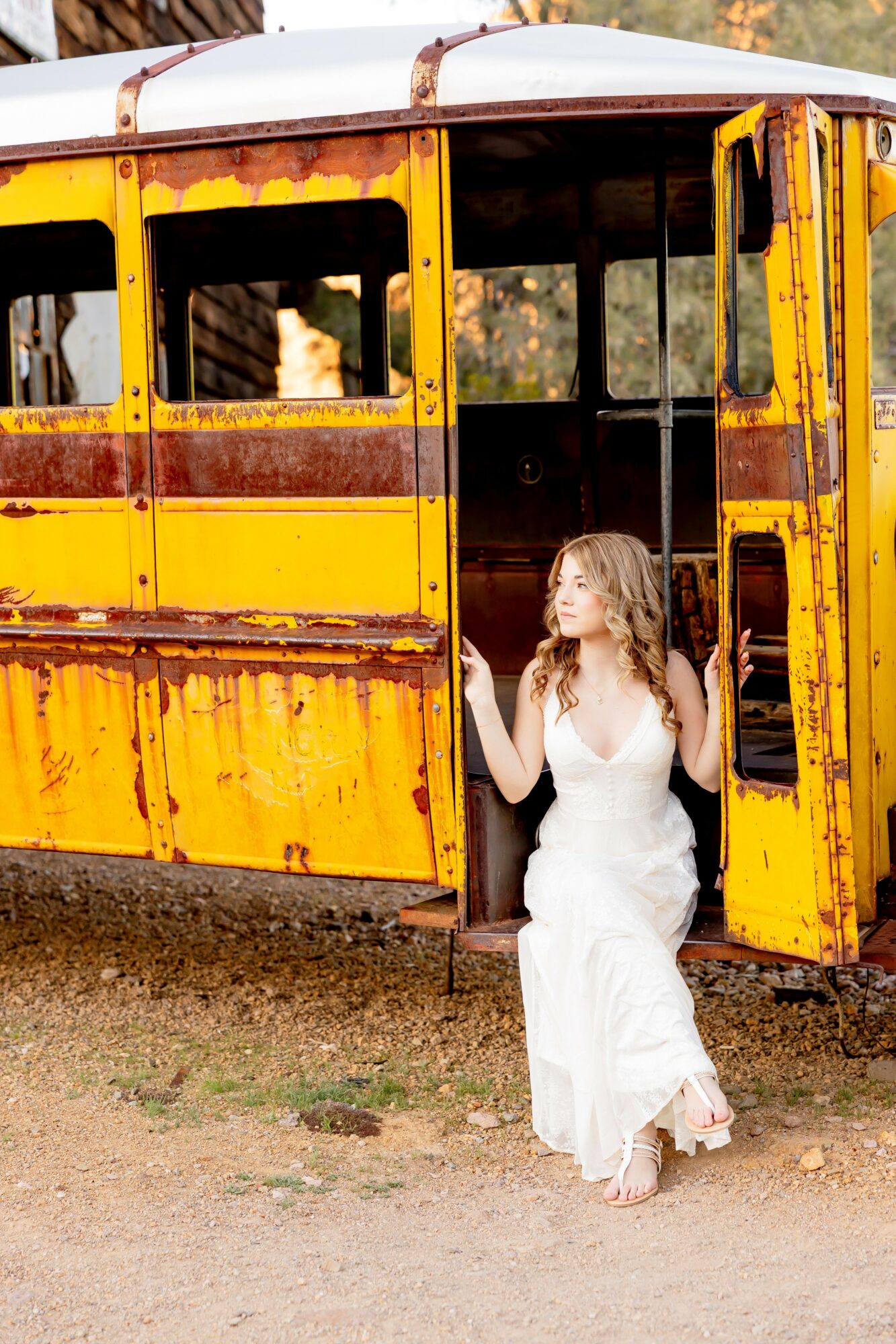 Young woman in white dress standing at open door of yellow, rusty train car outdoors.