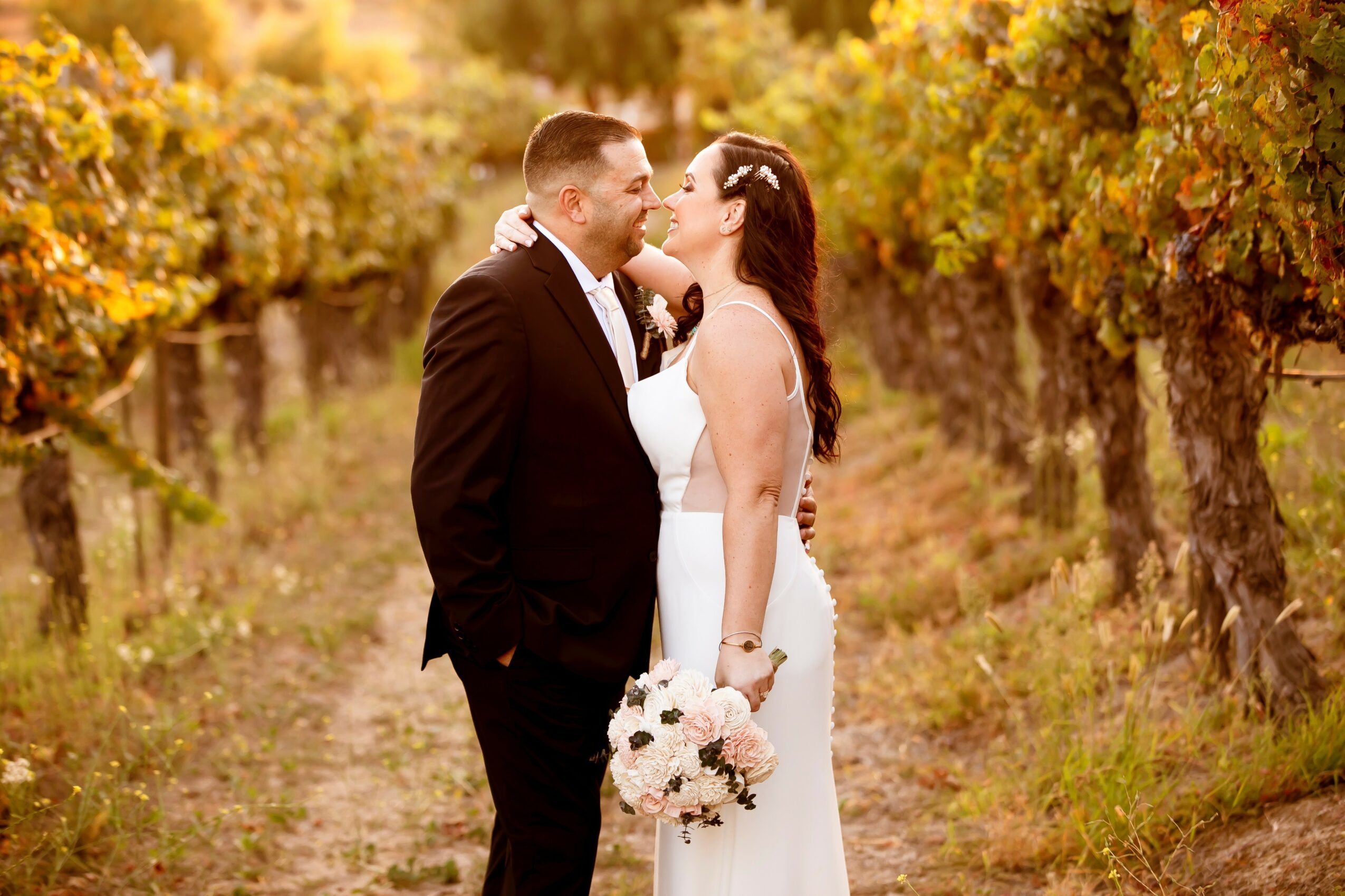 A bride and groom kiss in a vineyard with golden foliage, the bride holding a bouquet of flowers, both dressed formally.