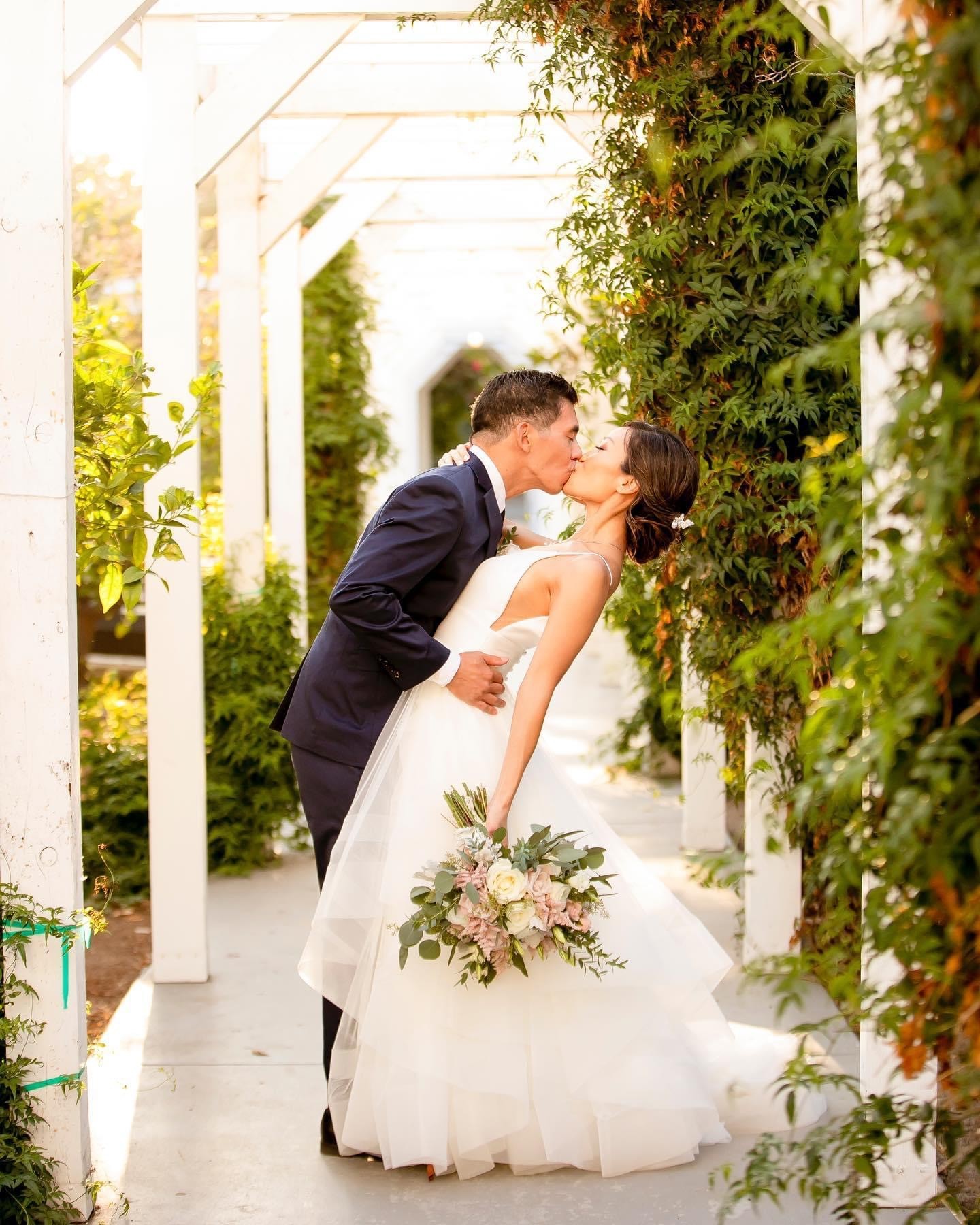 Couple in wedding attire sharing a kiss in a garden with greenery and white structures.