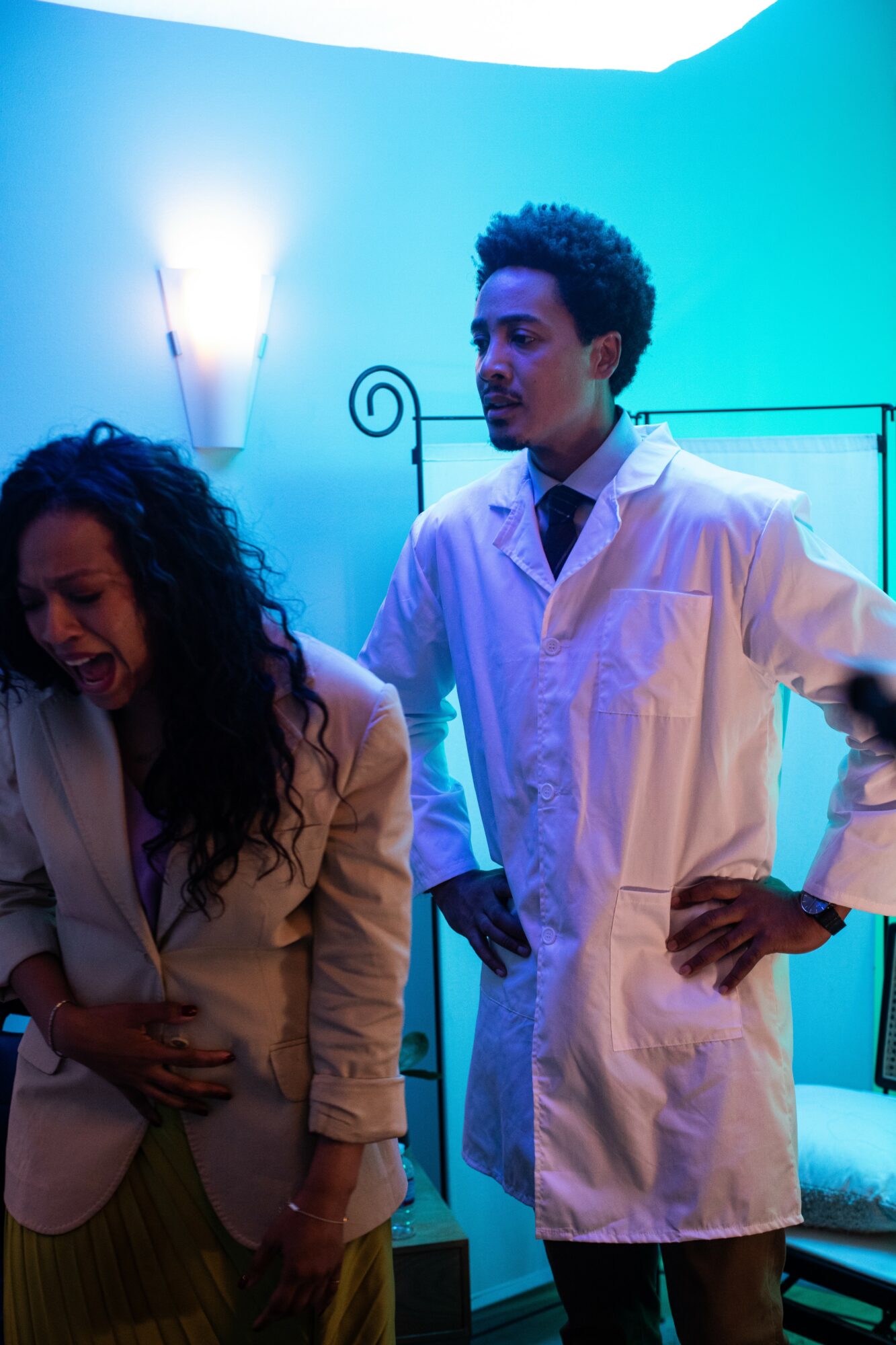 Two people in white lab coats stand in a room with blue lighting, one with hands on hips, the other looking down.