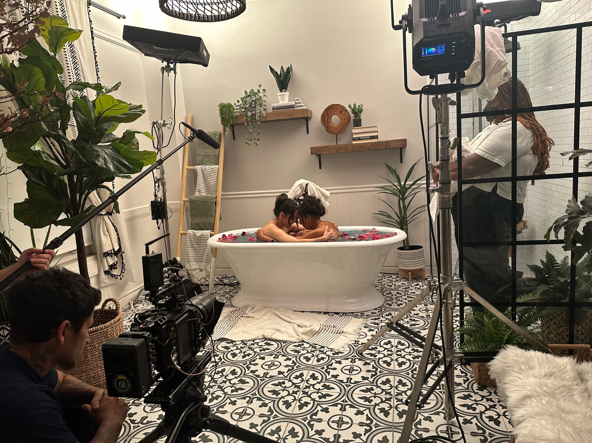Child in bathtub with adult, surrounded by filming equipment and crew, in a decorated room with plants and patterned floor.
