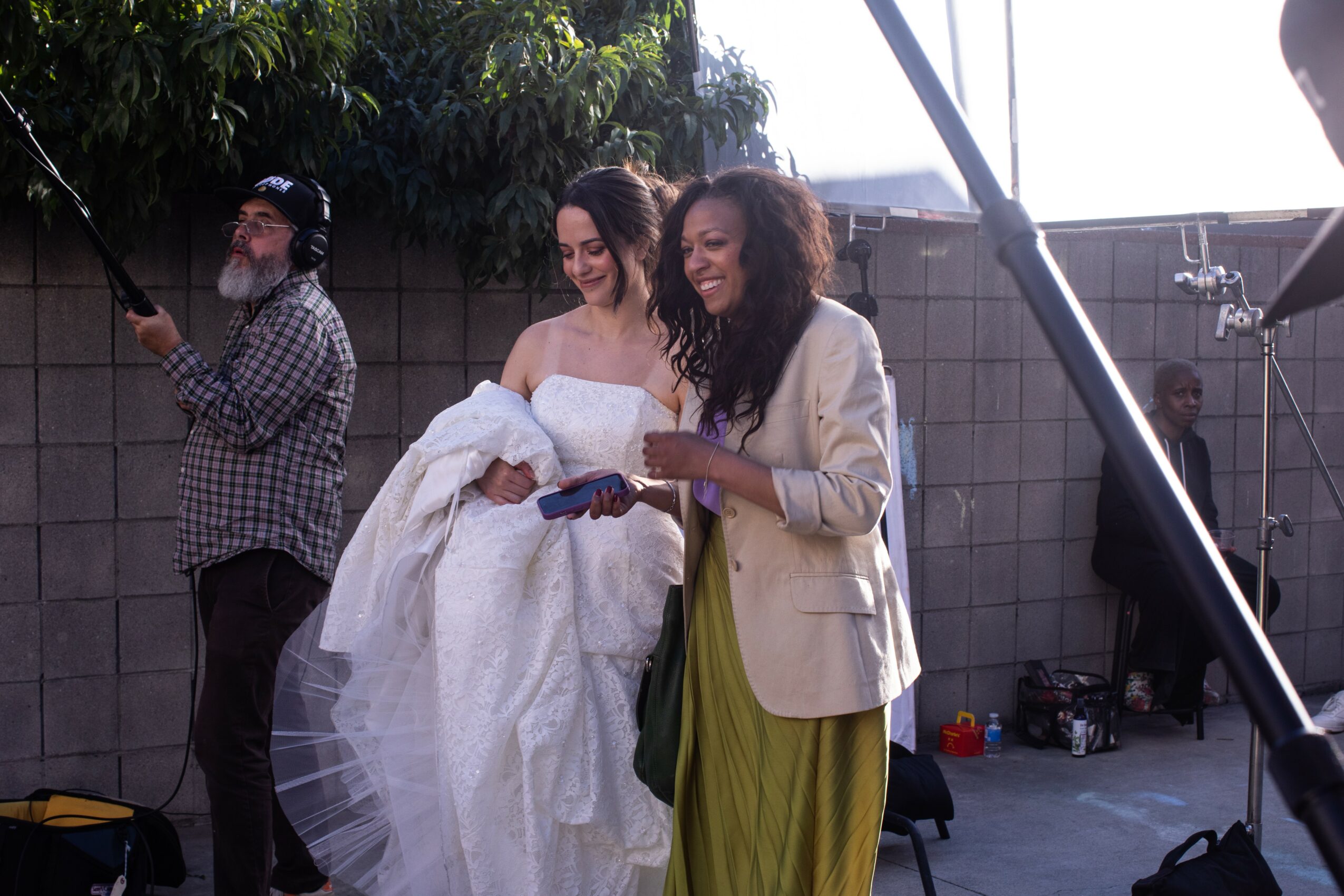 Two women in wedding attire smiling and looking at a phone, outdoors with a brick wall and trees in background.