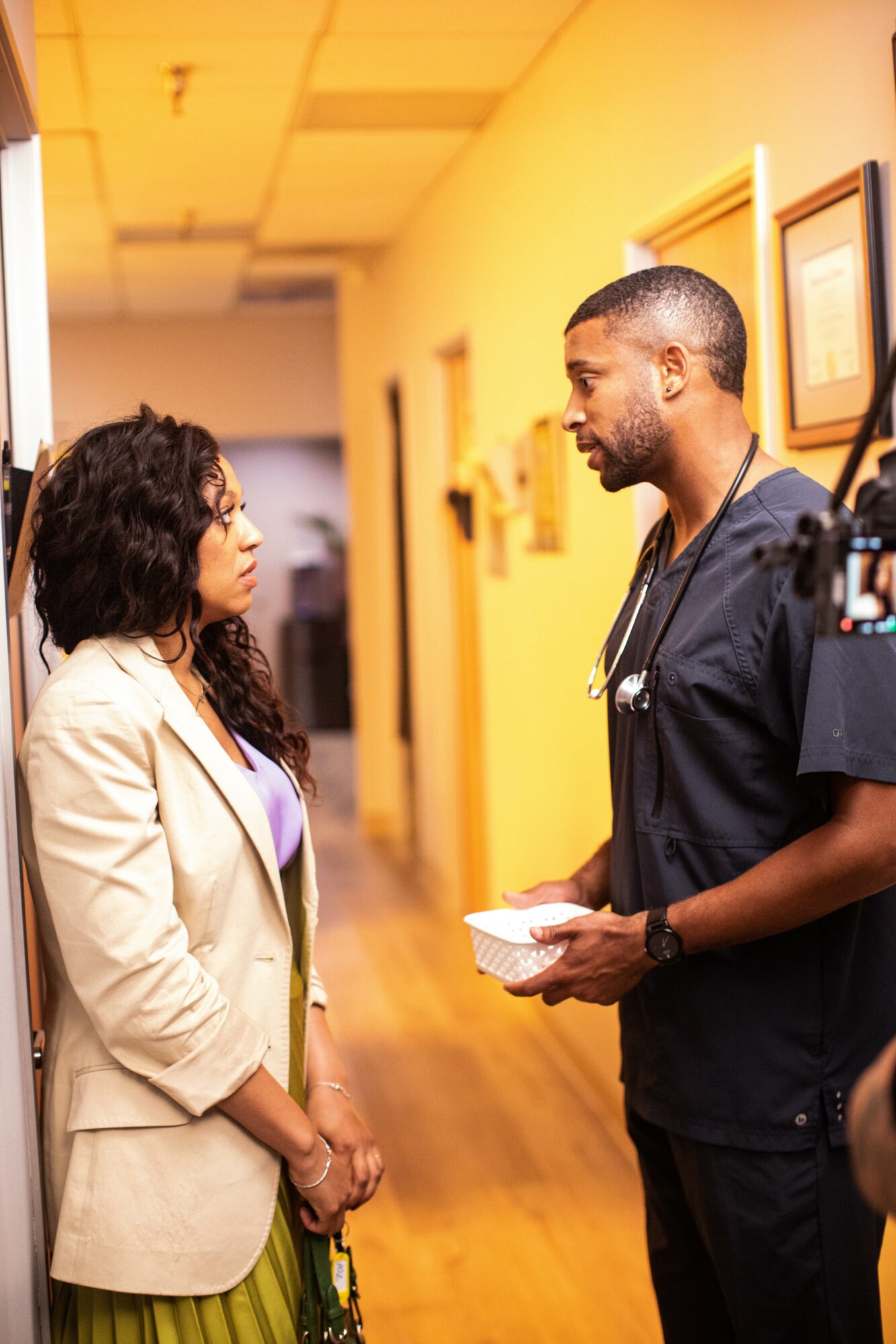A woman and a man, possibly a healthcare professional, are engaged in conversation in a hallway with framed pictures on the wall.