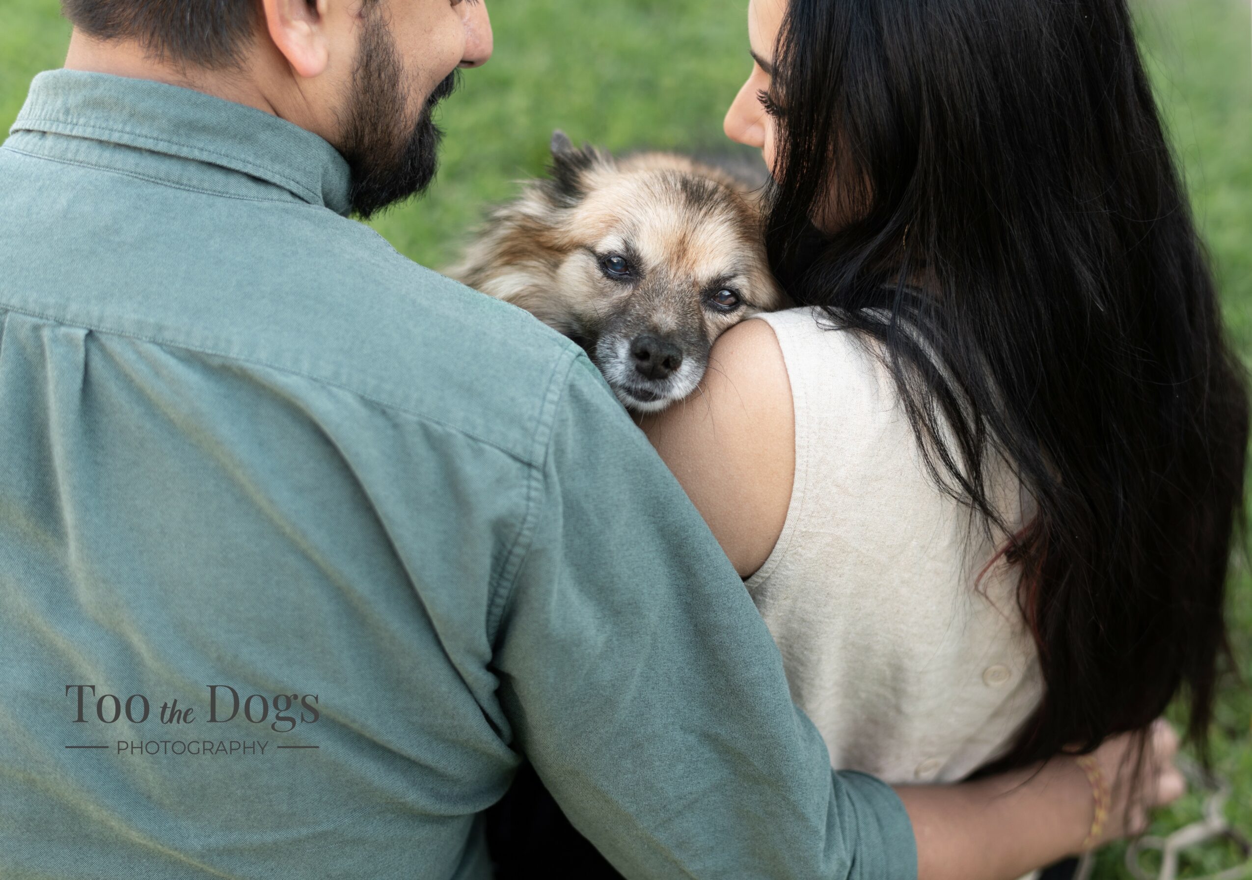 Two people hold a dog between them outdoors, with green grass in the background.