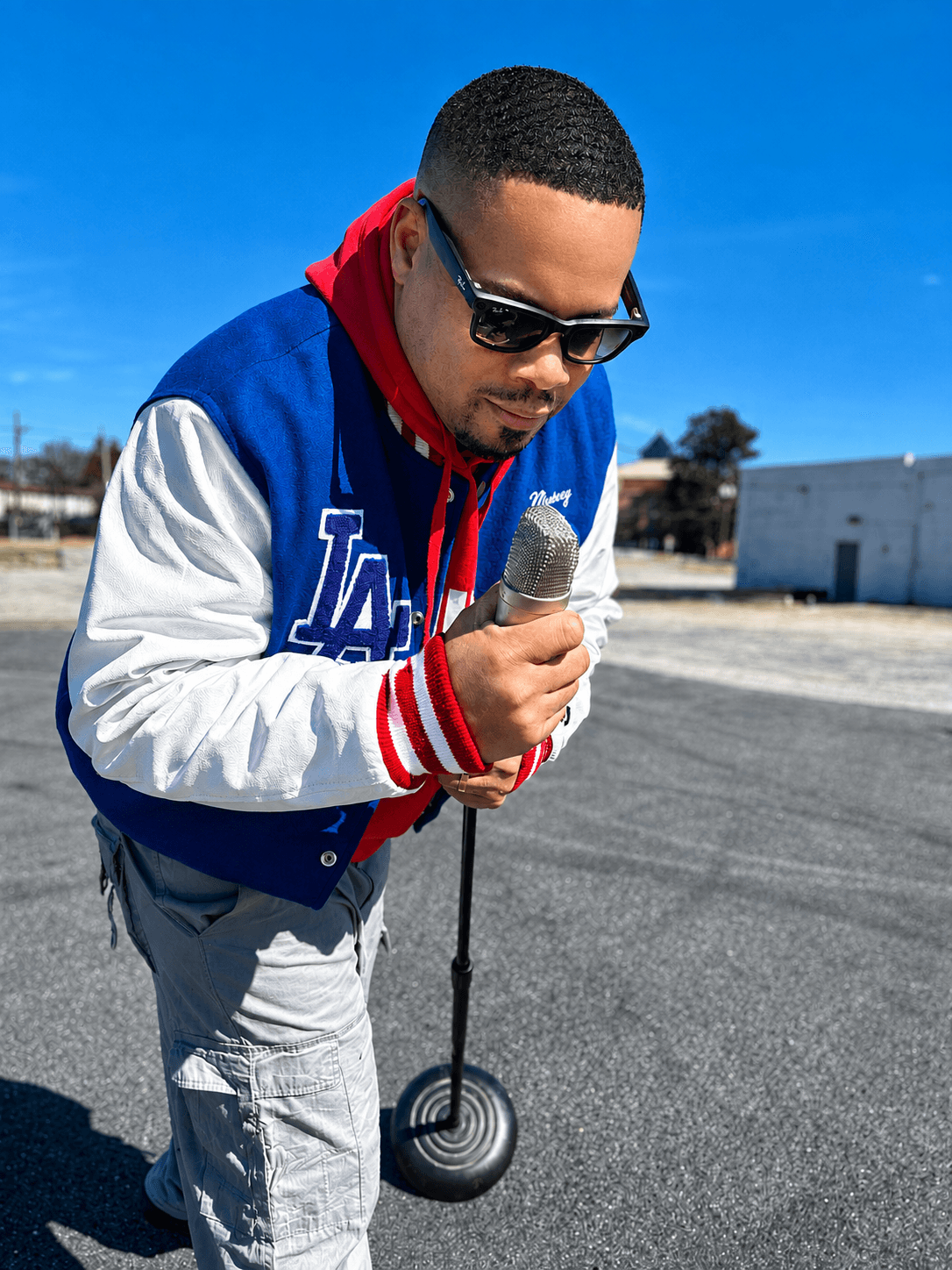 Person wearing sunglasses and a blue and white jacket, holding a microphone on a street, with a clear blue sky.