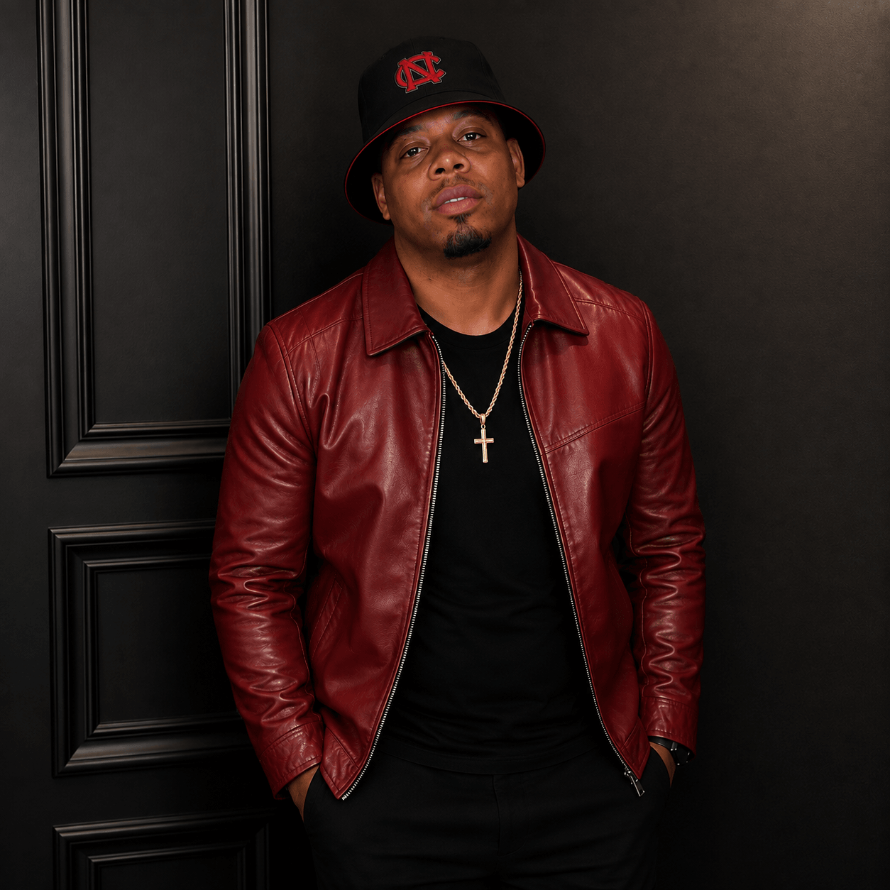 Man wearing a black hat with red logo, red leather jacket, black shirt, gold cross necklace, standing against black background.