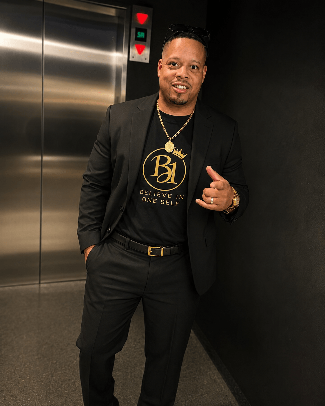 Man in black suit and T-shirt standing in elevator, smiling, pointing with his right hand, black wall background.