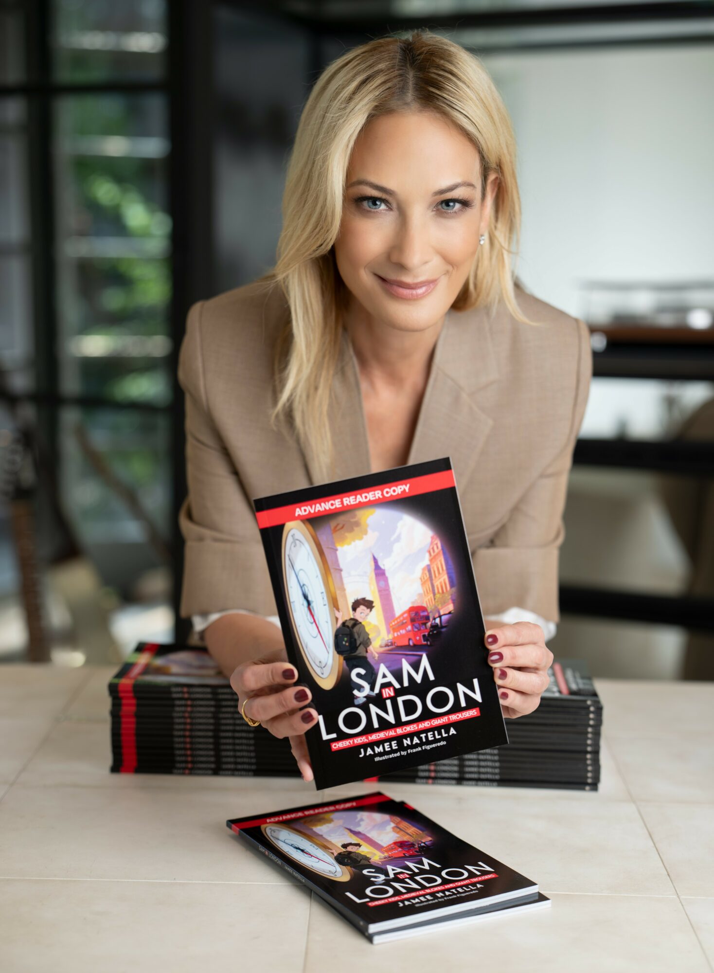 Woman with blonde hair holding a book titled 'Sam London' in a modern room, smiling at the camera.