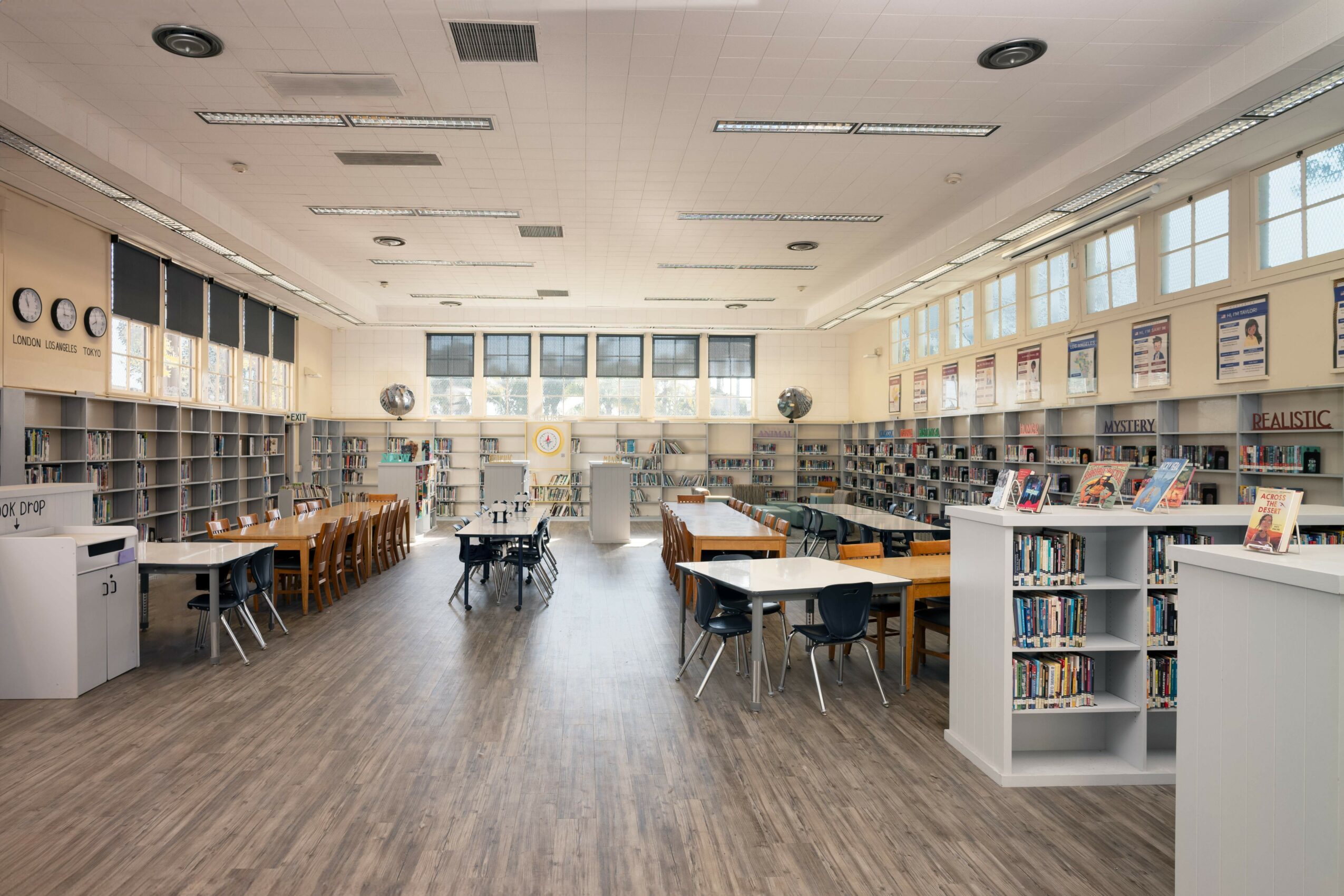 Empty library with bookshelves, tables, and chairs, large windows, and wooden flooring.