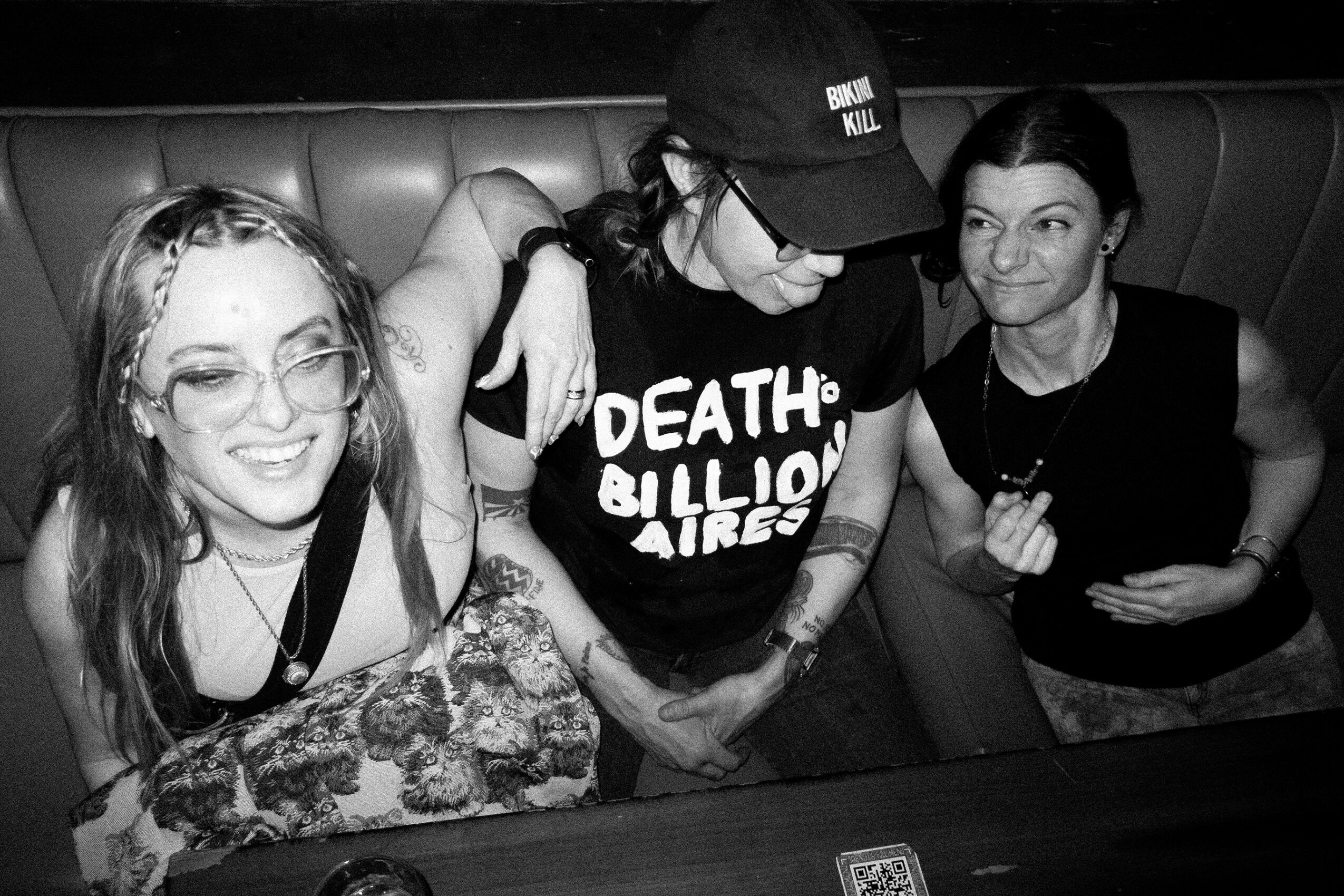 Three women sitting together at a table, smiling and engaging with each other, in a black-and-white photo.
