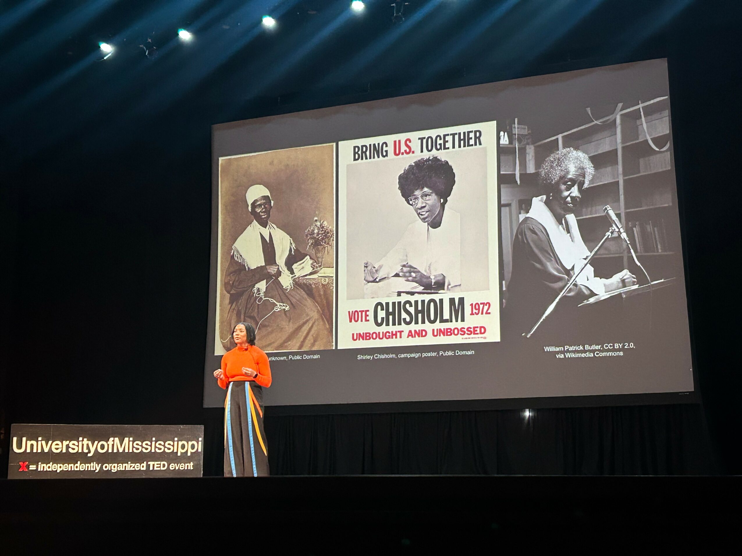 Person in orange shirt standing on stage with three posters about Chisholm and U.S. history projected behind them.