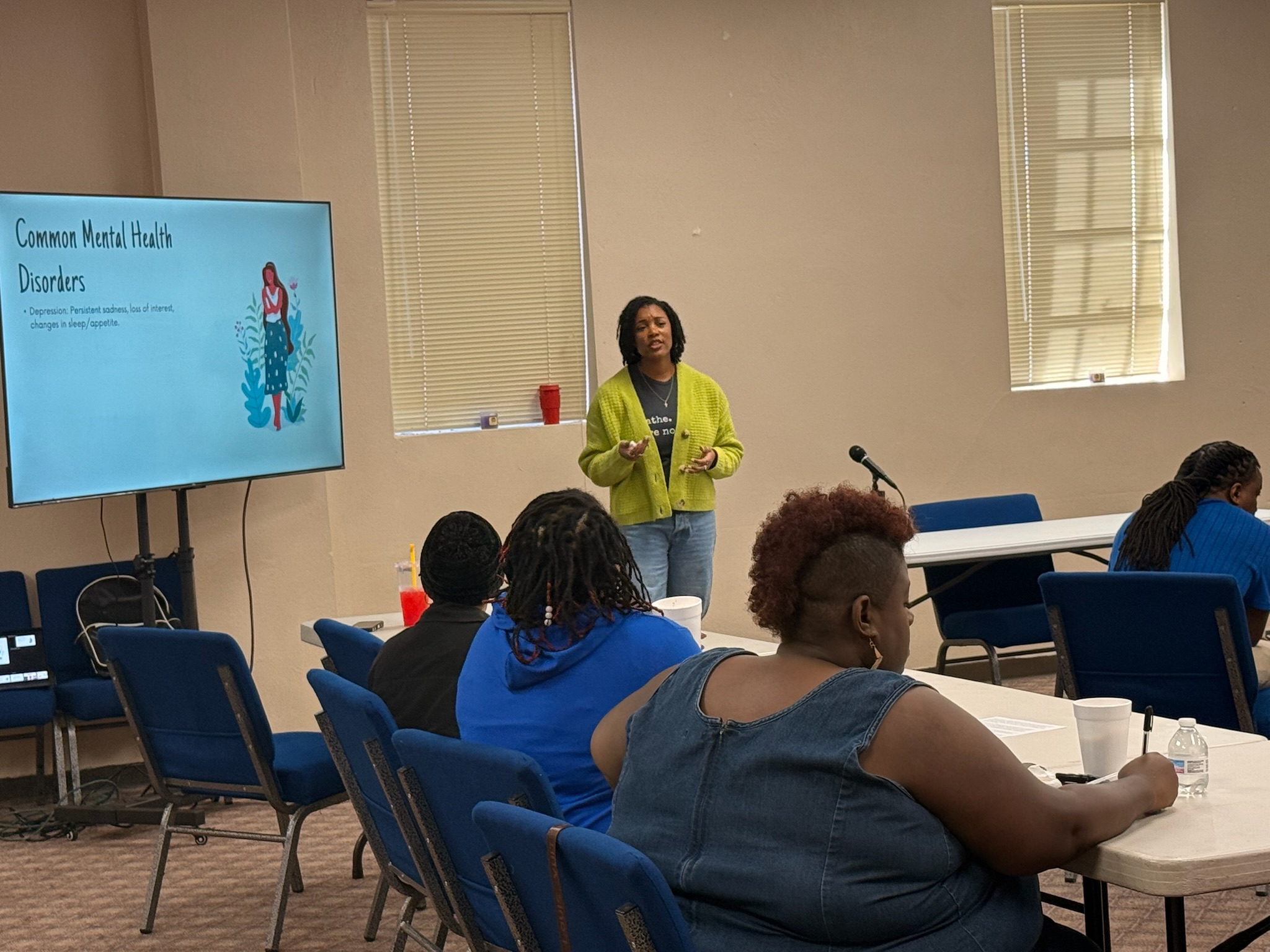 Woman standing and speaking in front of a group in a conference room with a large screen displaying a presentation.
