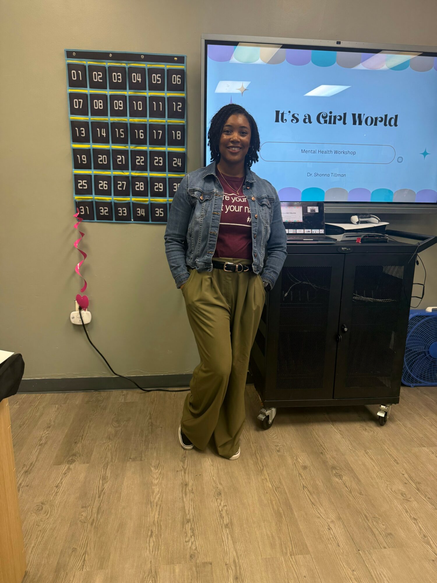 Woman standing in classroom with a colorful calendar and a screen displaying 'It's a Girl World'.