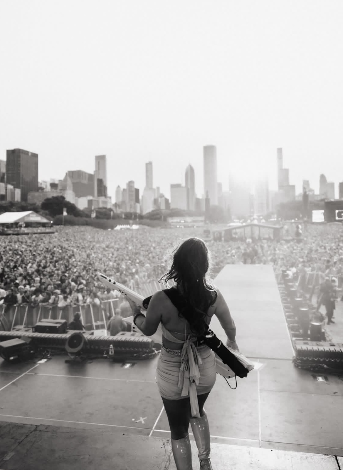 A woman stands on stage overlooking a large outdoor crowd with a city skyline in the background.