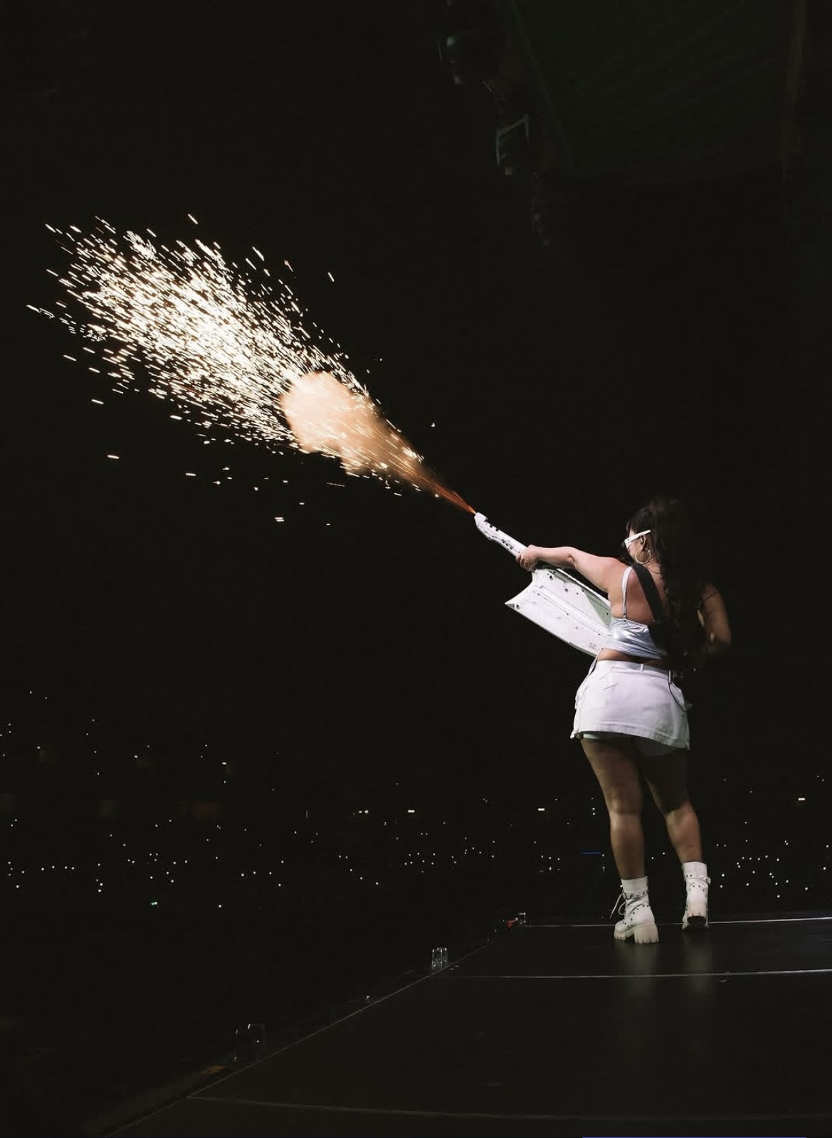 Person in white dress holding a firework that sprays sparks into the air, dark background, stage setting.