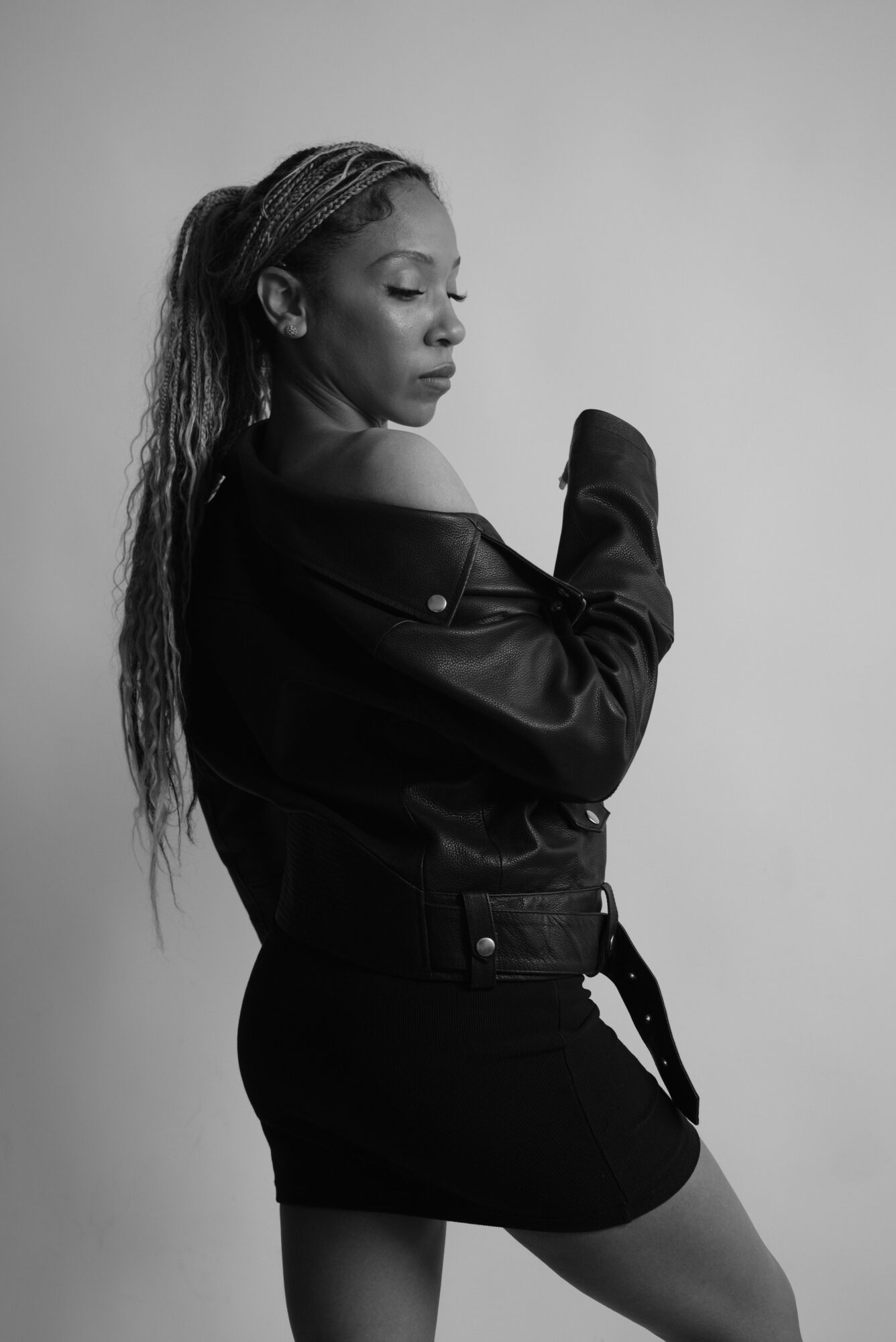 A woman with long hair wearing a leather jacket and shorts, standing sideways against a plain background.