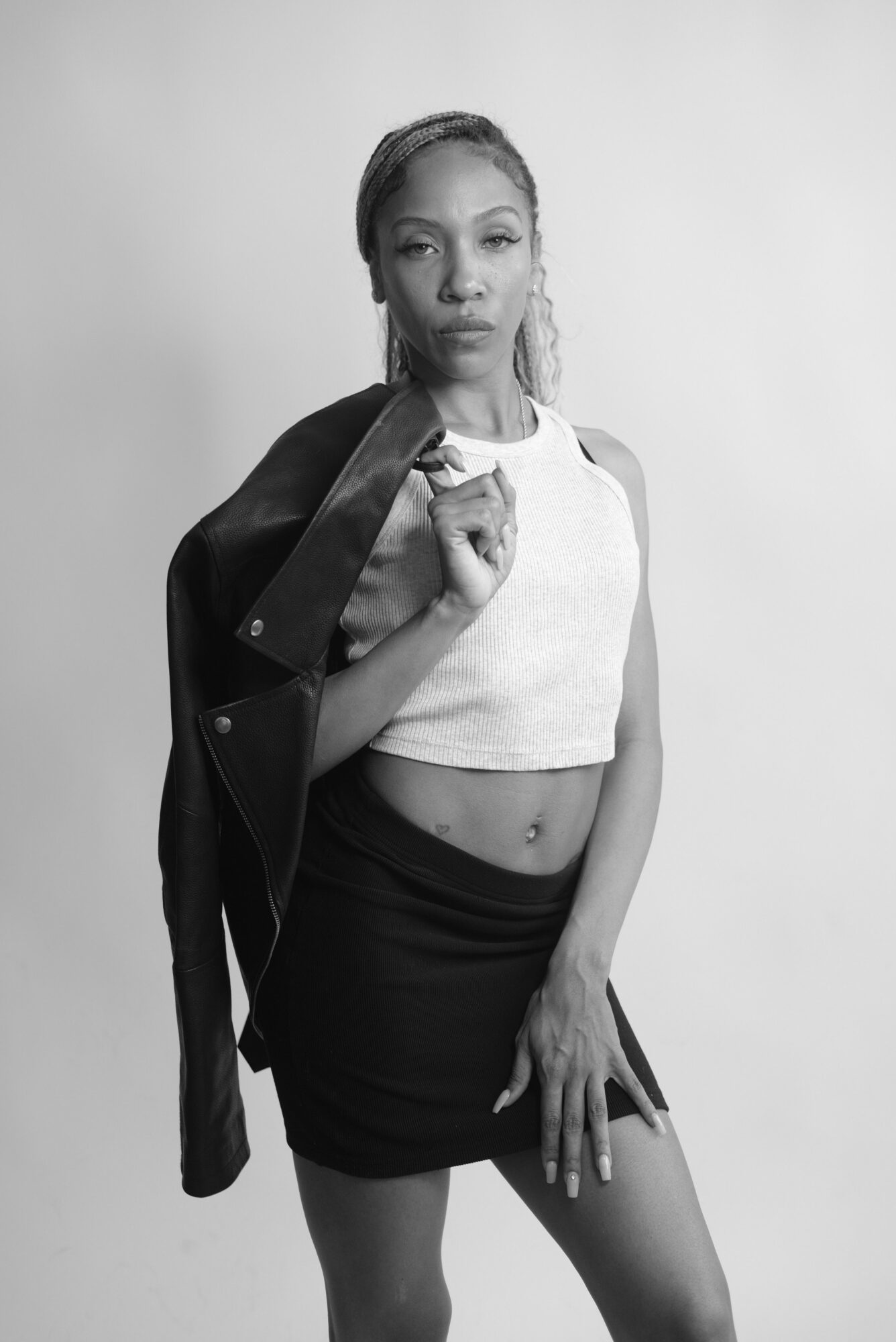 A woman with a leather jacket draped over her shoulder, wearing a white top and black shorts, standing against a plain background.