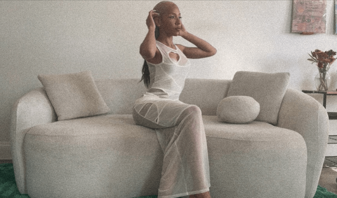 Person sitting on a light-colored sofa, adjusting their hair, in a room with a white wall and a small table with a plant.