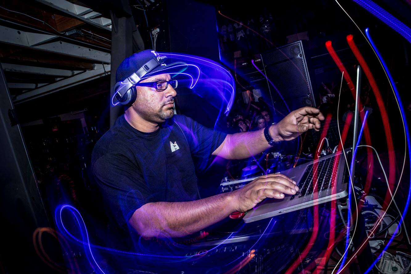 DJ wearing headphones and glasses, working with electronic equipment in a dark, colorful setting.