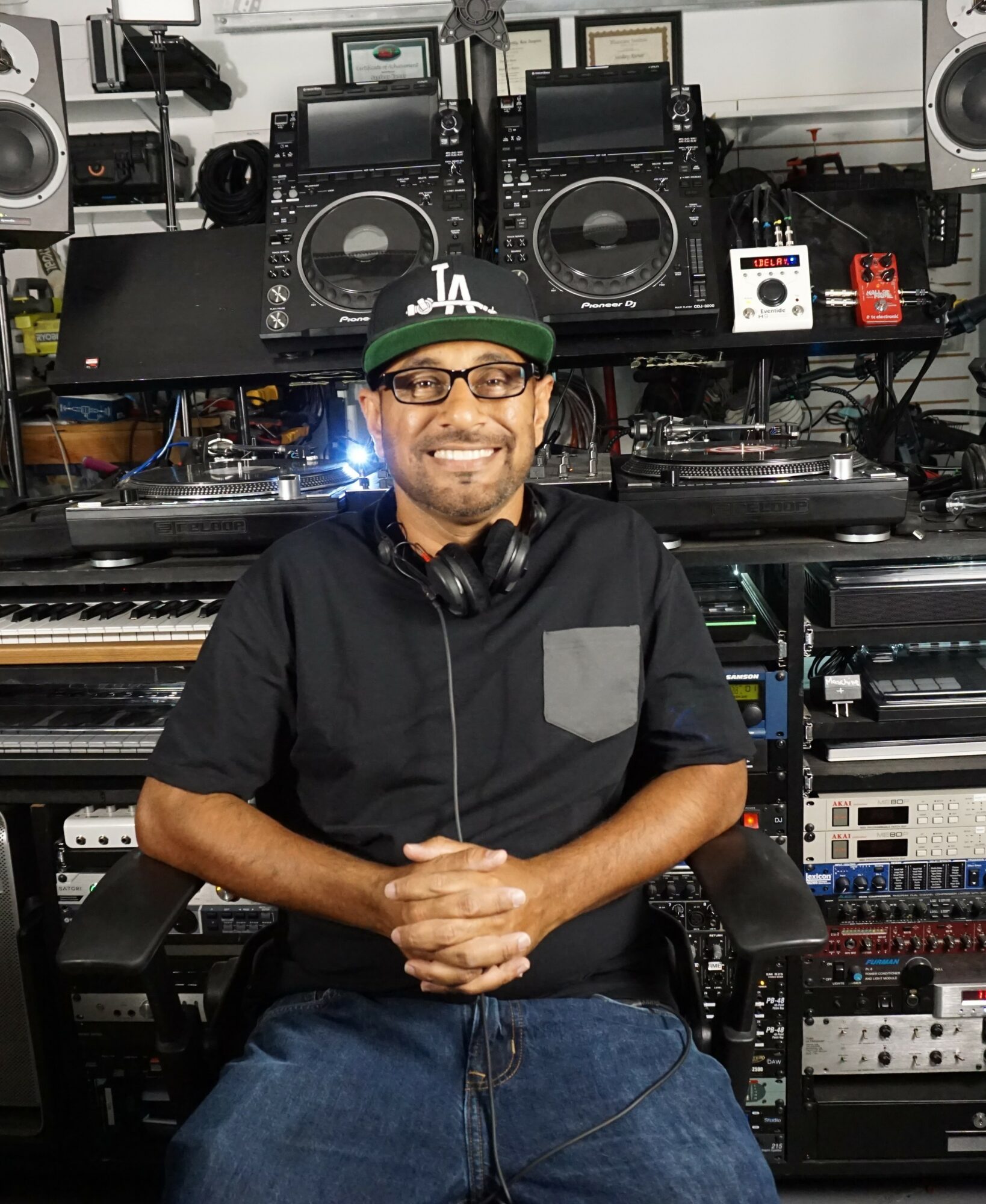 Man with glasses and a cap sitting in a music studio with DJ equipment and speakers behind him.