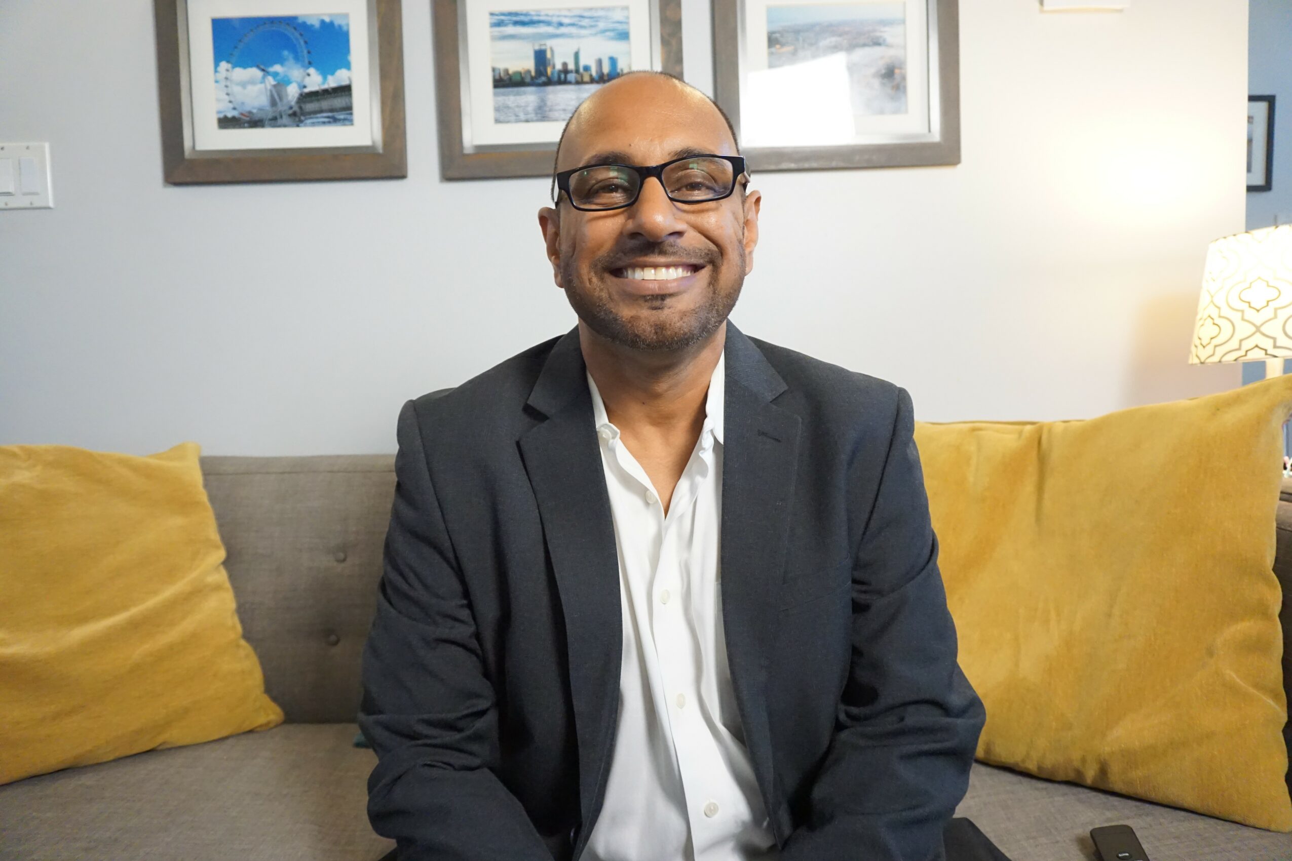 Man with glasses in a suit smiling, sitting on a sofa with yellow pillows, framed pictures on the wall behind.