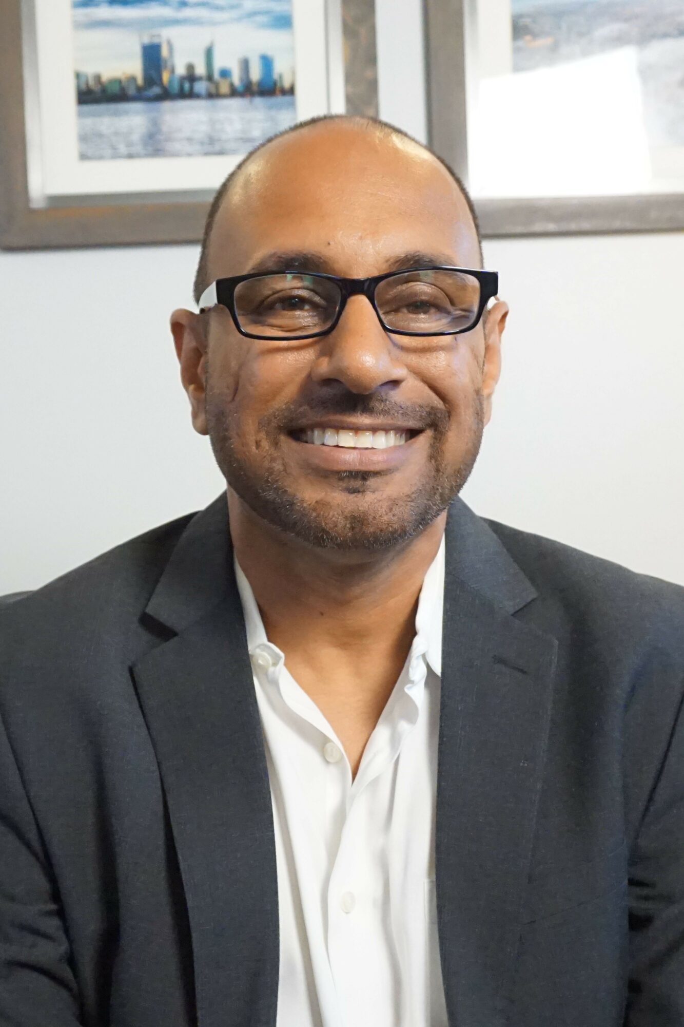 Man wearing glasses and a suit, smiling, in an office with a window and cityscape view