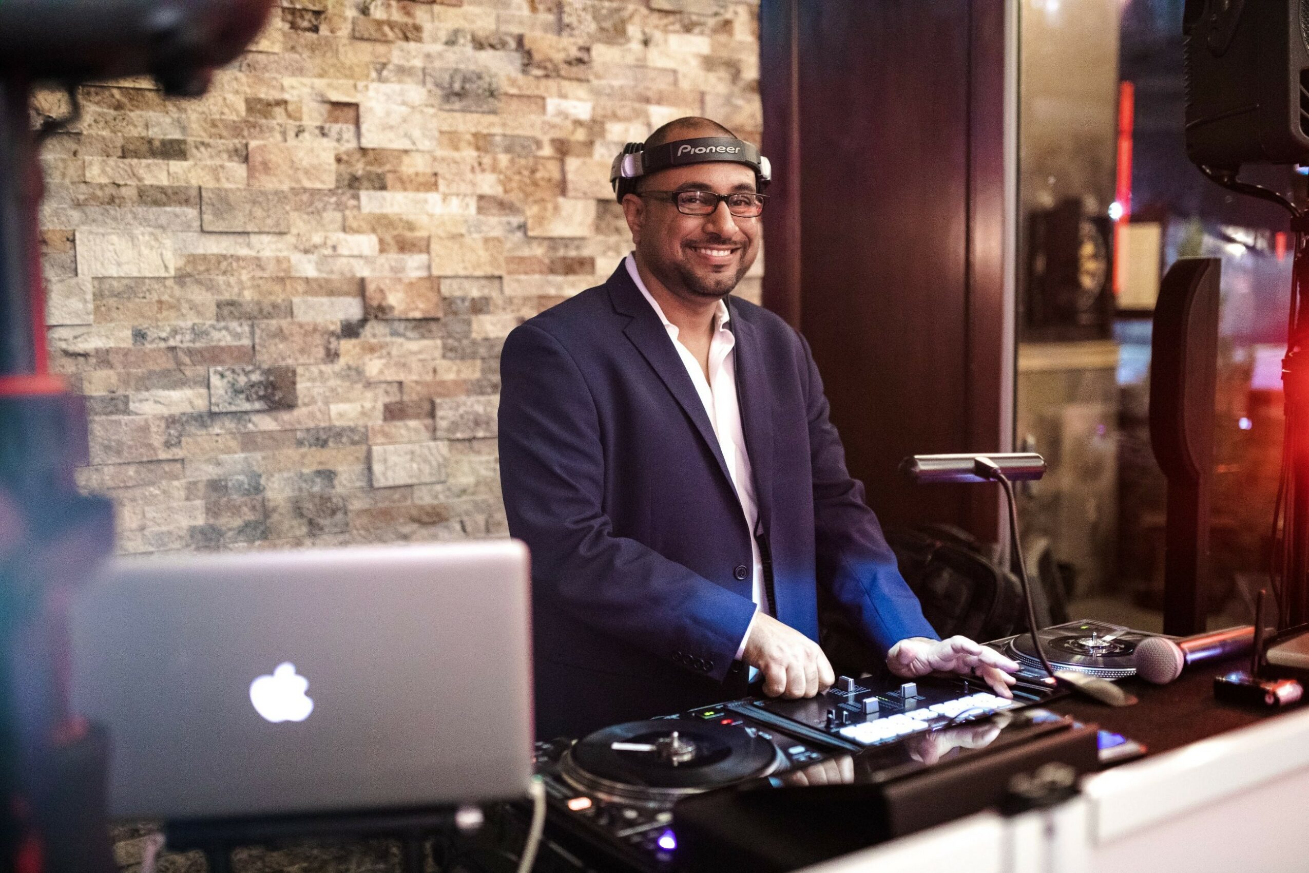 Man in suit DJing with equipment, smiling, in front of a stone wall, at an indoor event.