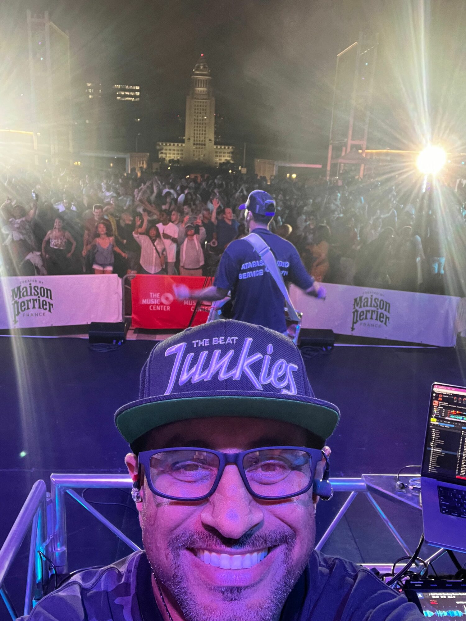 Person wearing glasses and a cap with 'Twinkies' logo, smiling at a DJ booth with a crowd and city skyline in background.