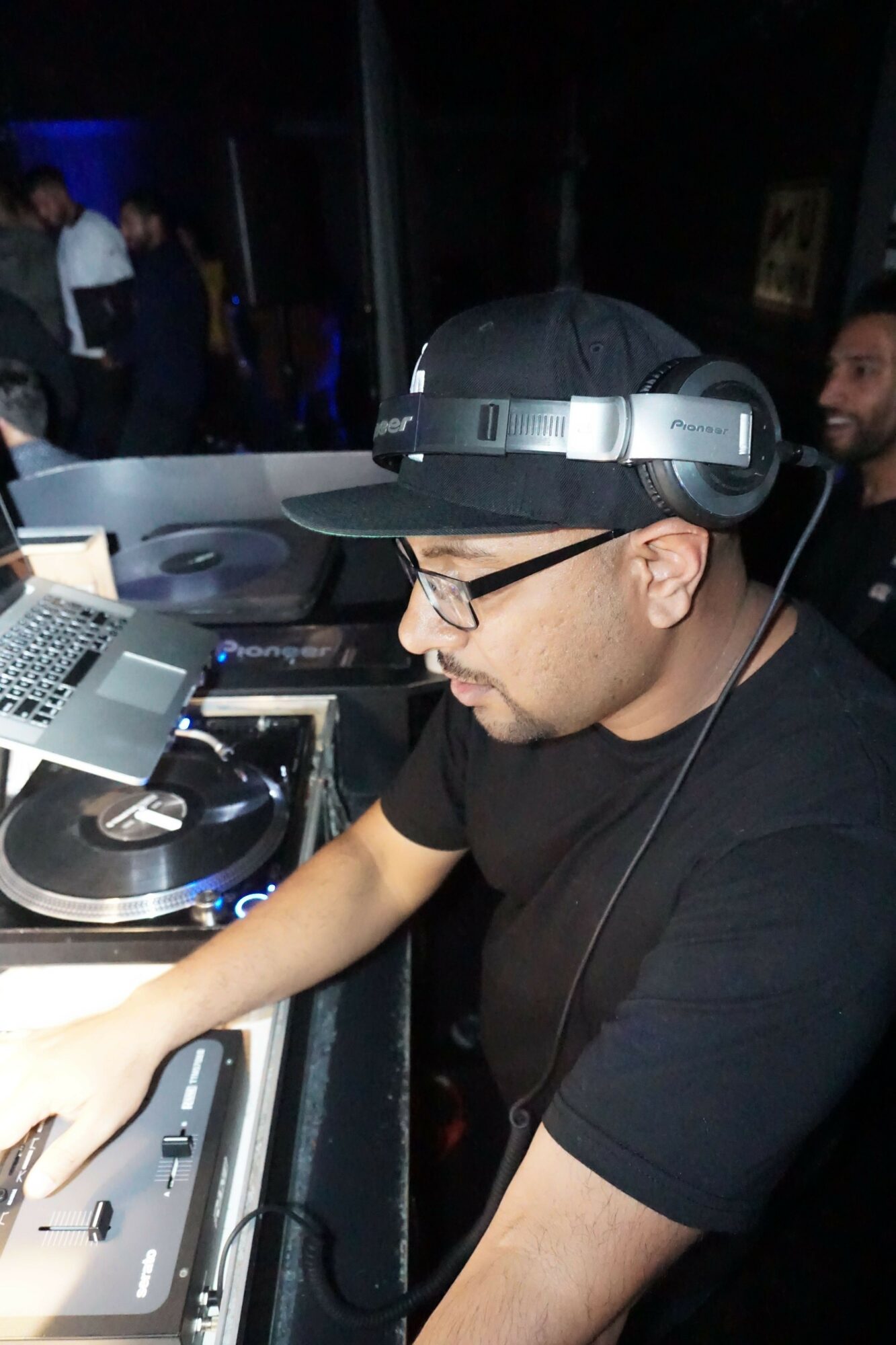 Person wearing headphones and glasses, DJing with equipment, in a dark environment with people in the background.