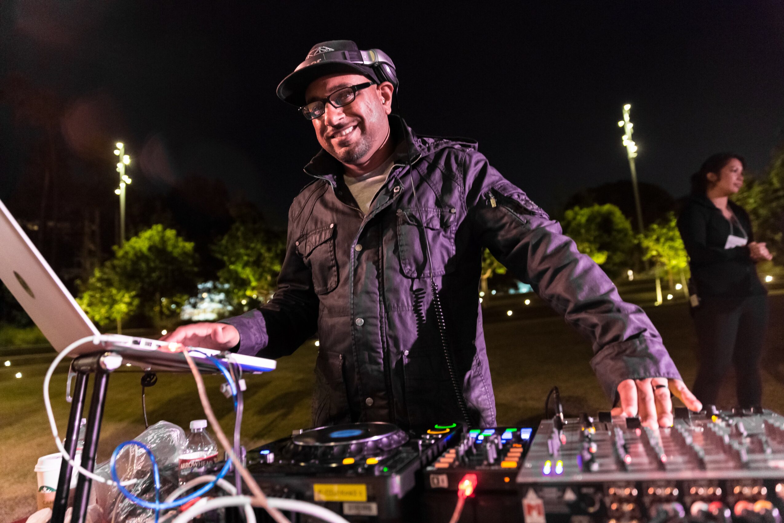 Man wearing glasses and a hat, smiling, operating DJ equipment outdoors at night, with trees and lights in background.