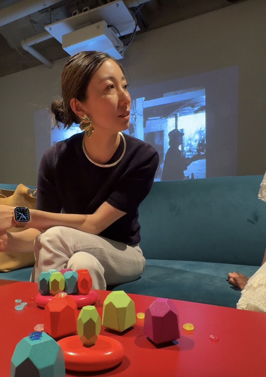 Woman sitting on a teal couch with colorful geometric toys on a red table in front, a projection on the wall behind her.