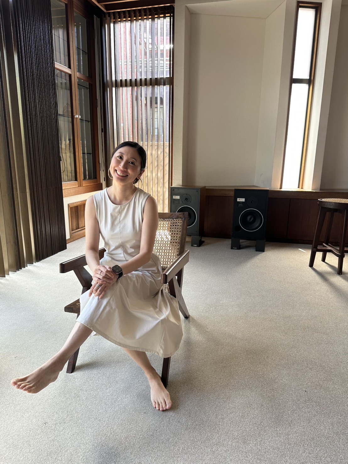 Woman in a white dress sitting on a chair indoors, with large windows and speakers in the background.