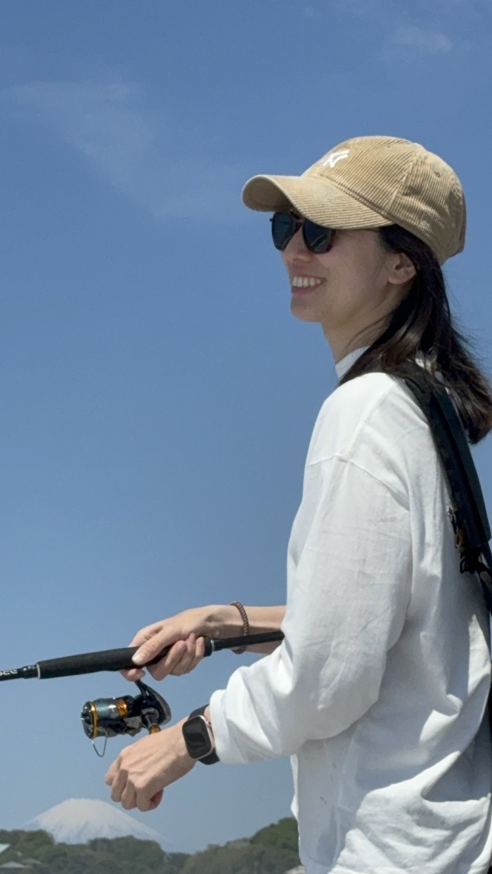 Woman wearing a beige cap and sunglasses, smiling, holding a fishing rod outdoors against a blue sky.