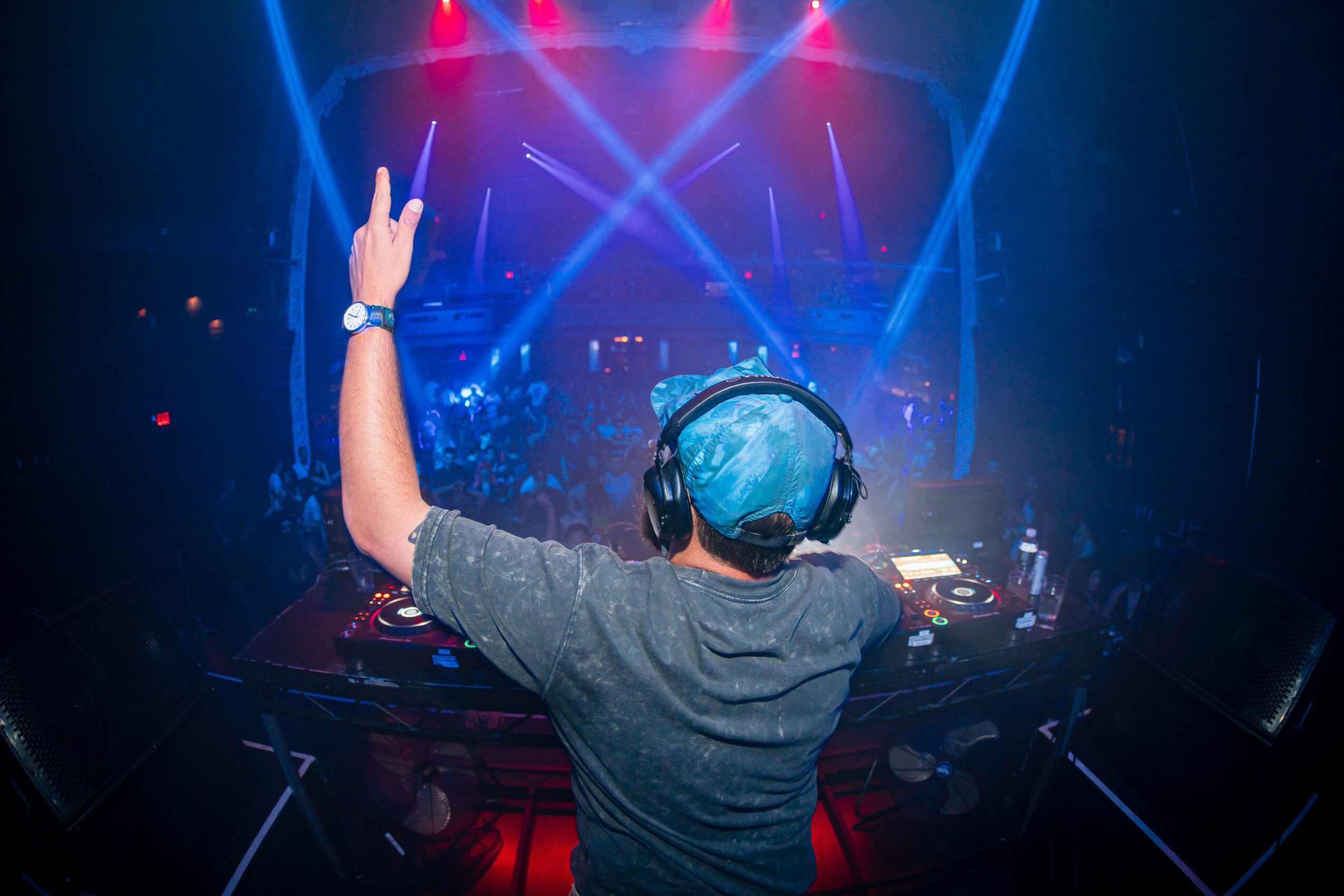 DJ wearing headphones and raising one hand in a dark, colorful concert or club setting with lights and equipment