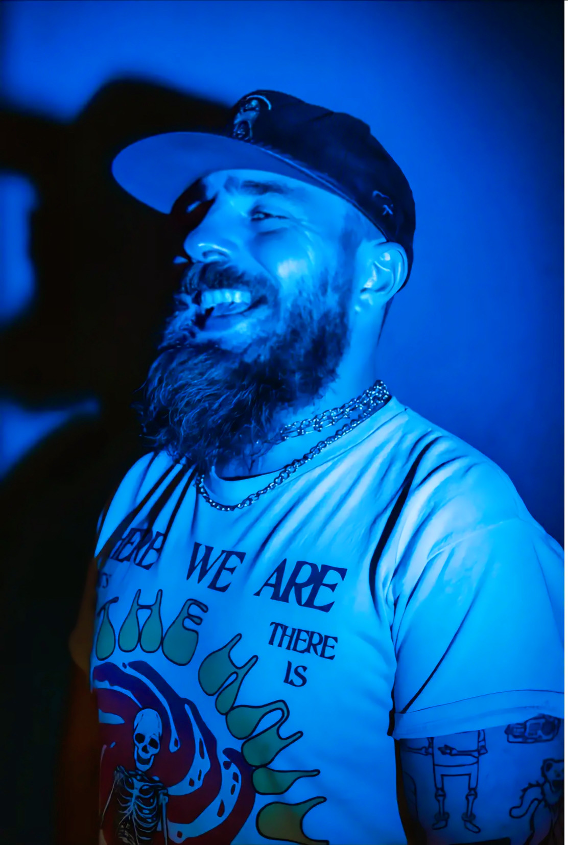 Smiling man with a beard, wearing a cap and a graphic T-shirt, illuminated by blue light.