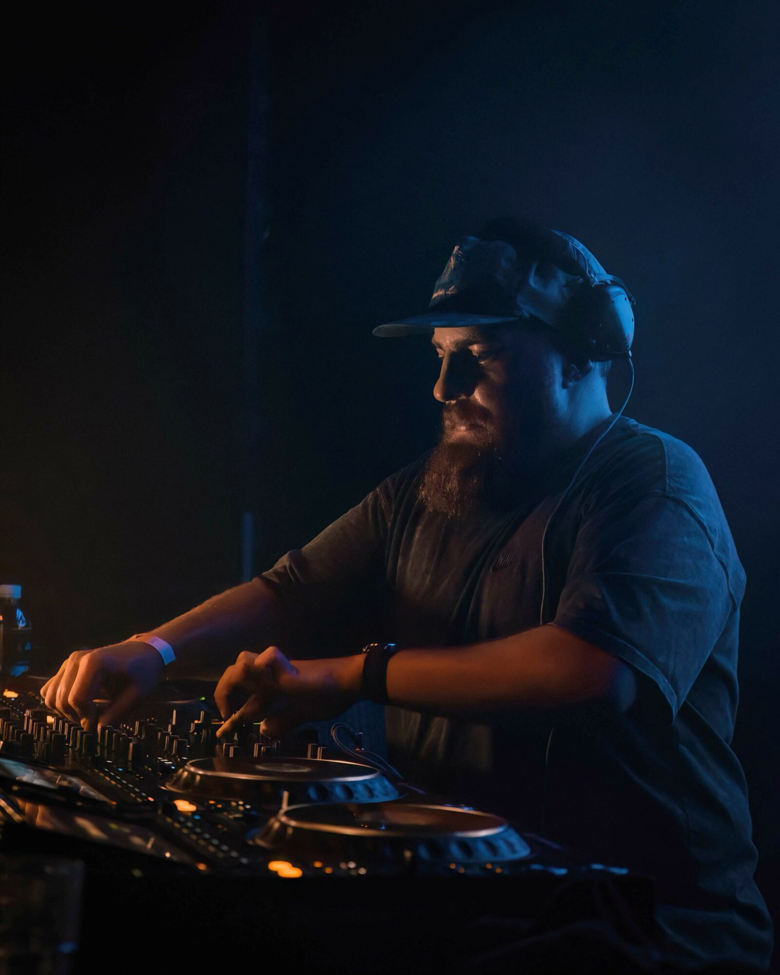 DJ with headphones operating turntables in dark setting, wearing sunglasses and a cap, focused on music equipment.