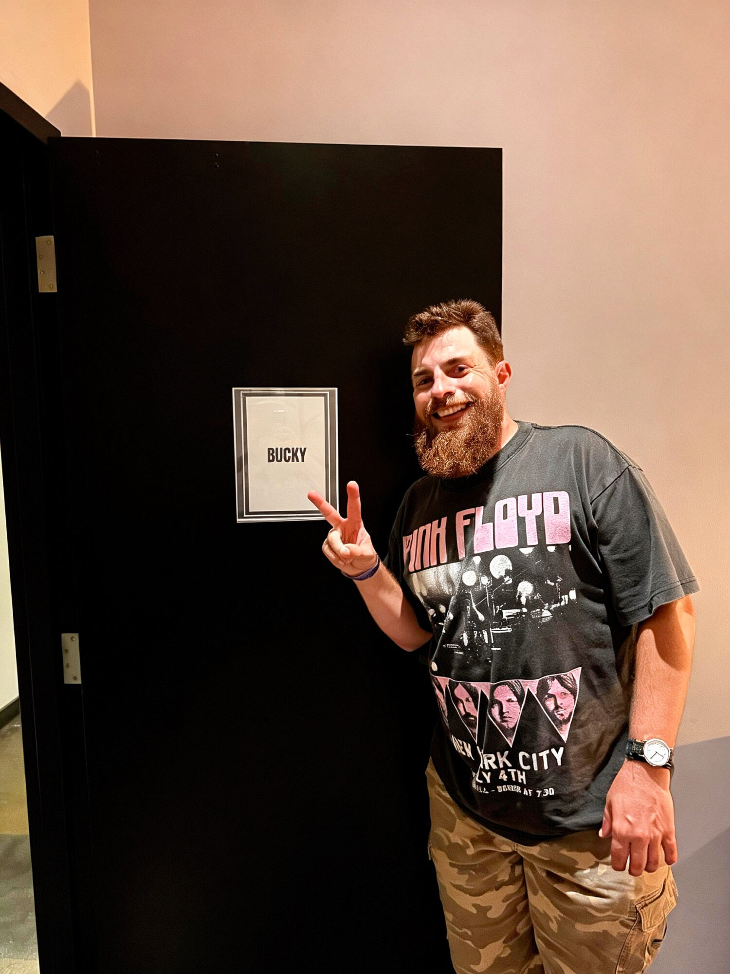 Man with a beard smiling and making a peace sign, standing next to a door with a sign that says 'BUCKY'.
