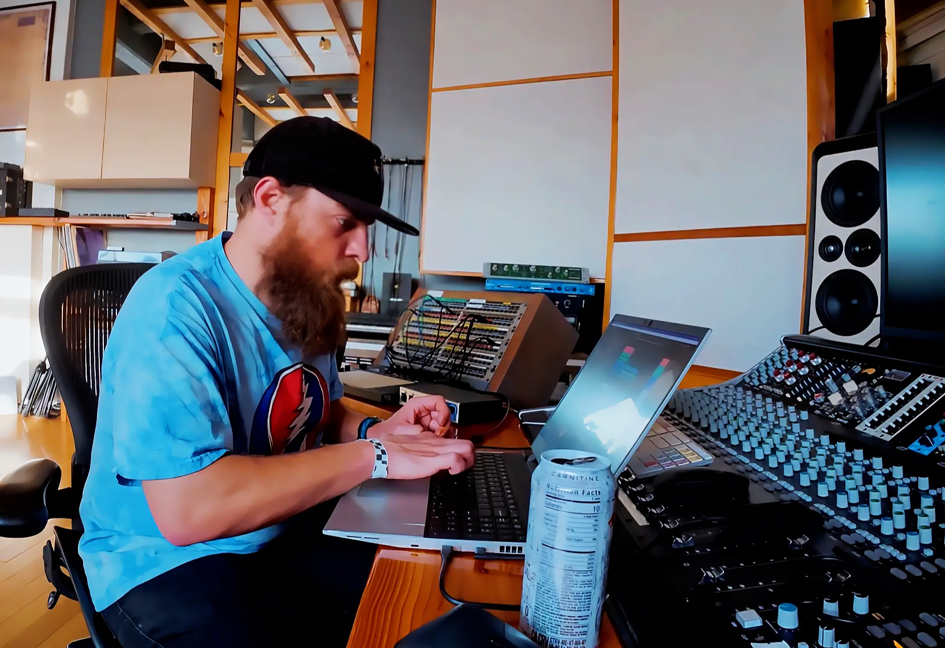 Person with a beard and cap working on a laptop in a music studio with audio equipment.