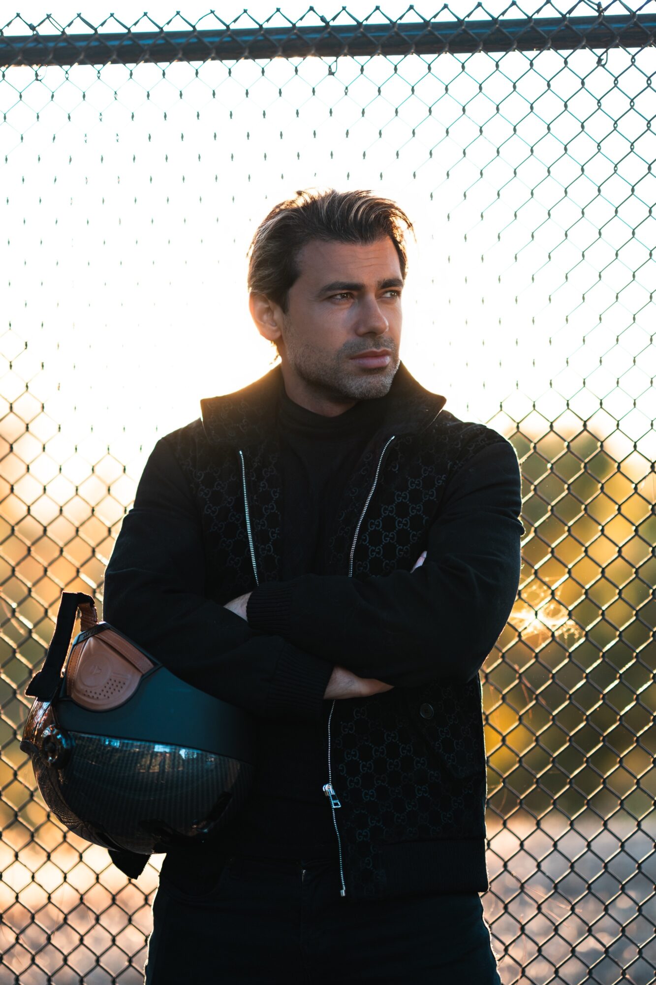 Man with dark hair and beard standing with arms crossed, holding a helmet, outdoors near a chain-link fence, sunset background.